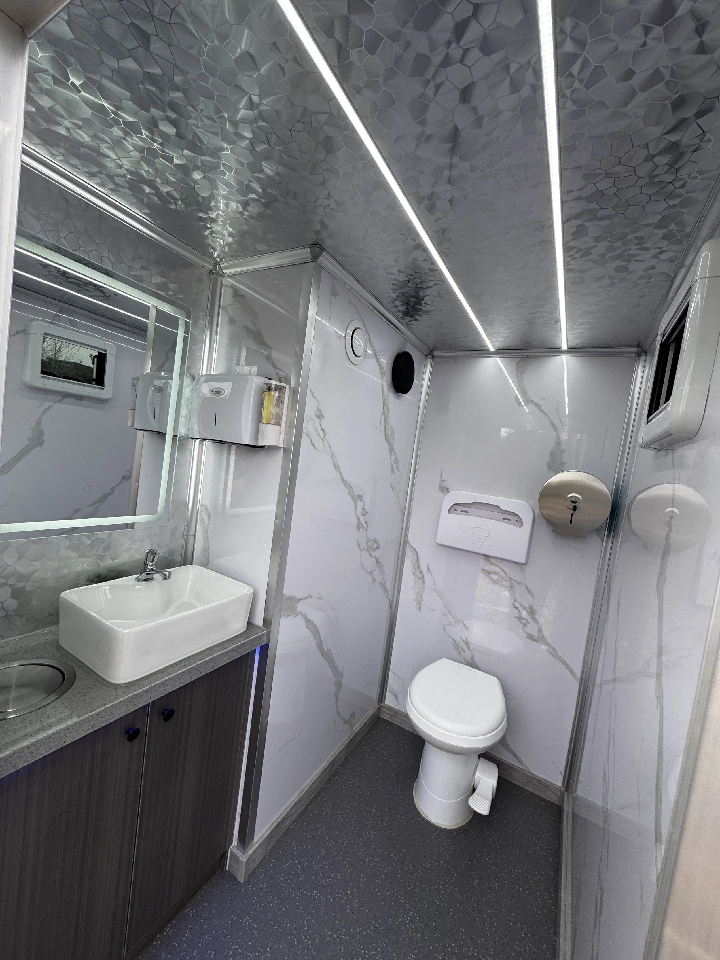 Small, modern bathroom with white marble walls, a toilet, a sink with a mirror, and medical supplies on the wall.