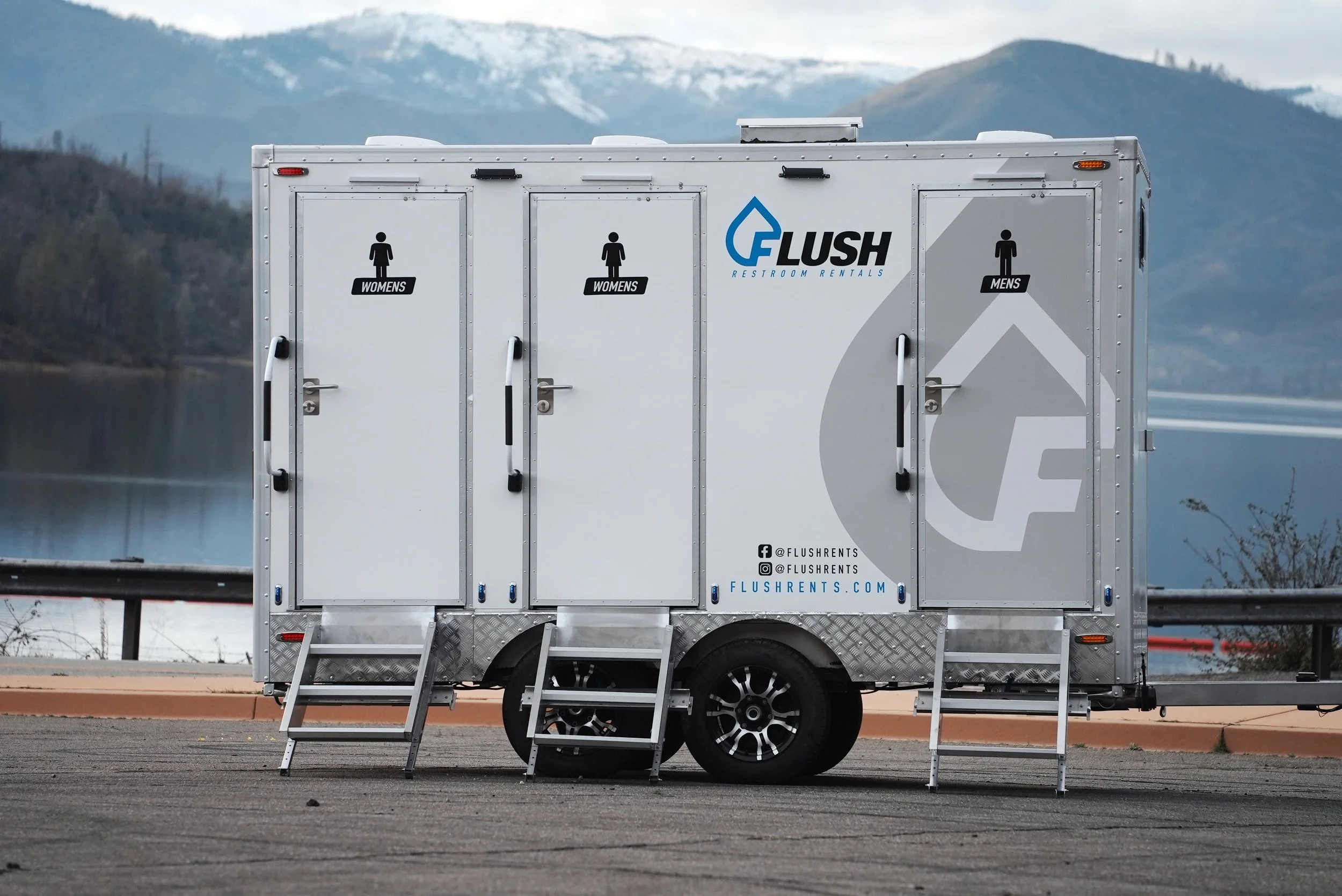 Portable restroom trailer with separate men's and women's entrances parked outdoors with mountains in the background.