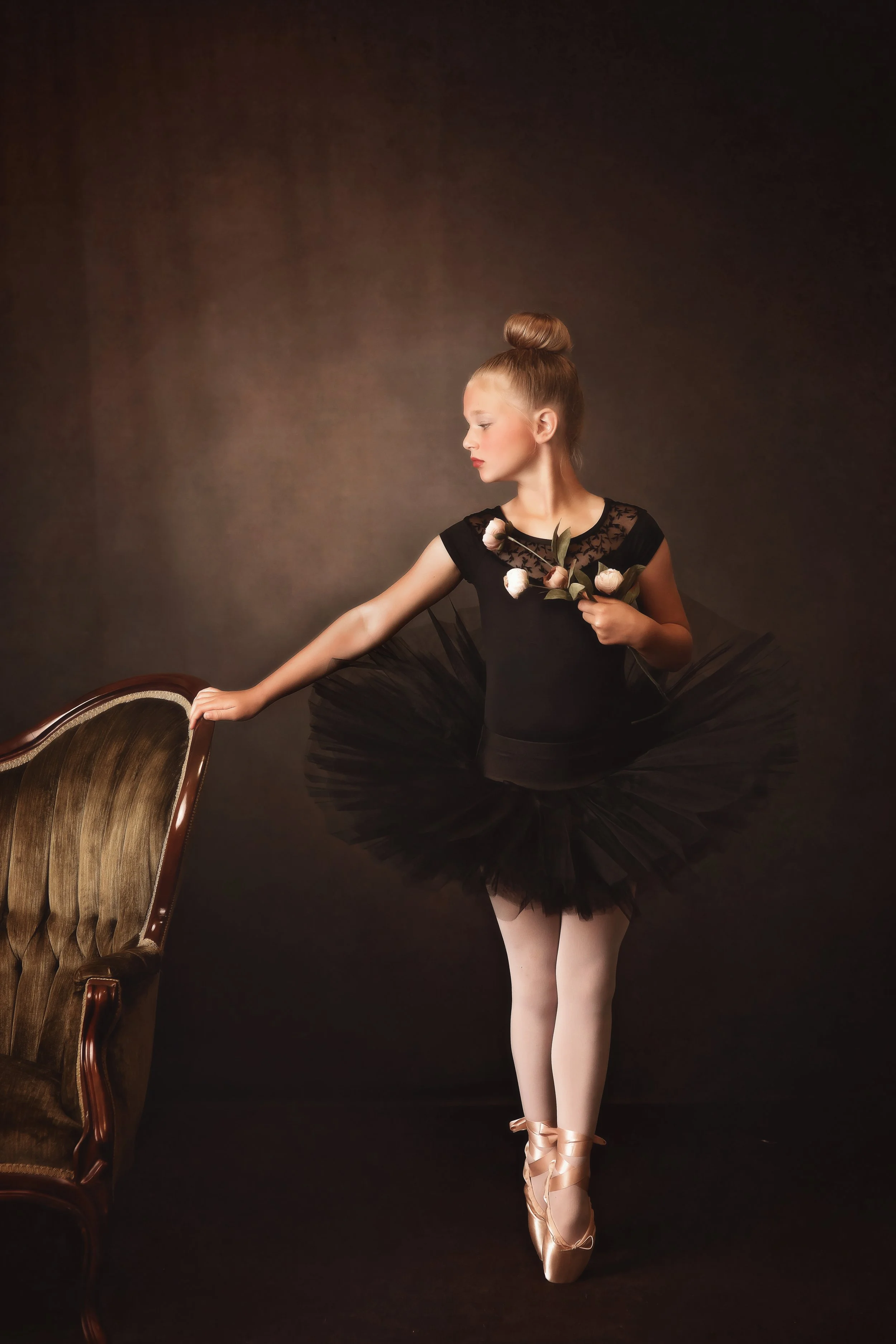 A ballerina in a black tutu, holding flowers and standing on pointe shoes, poses next to a vintage chair against a dark background.