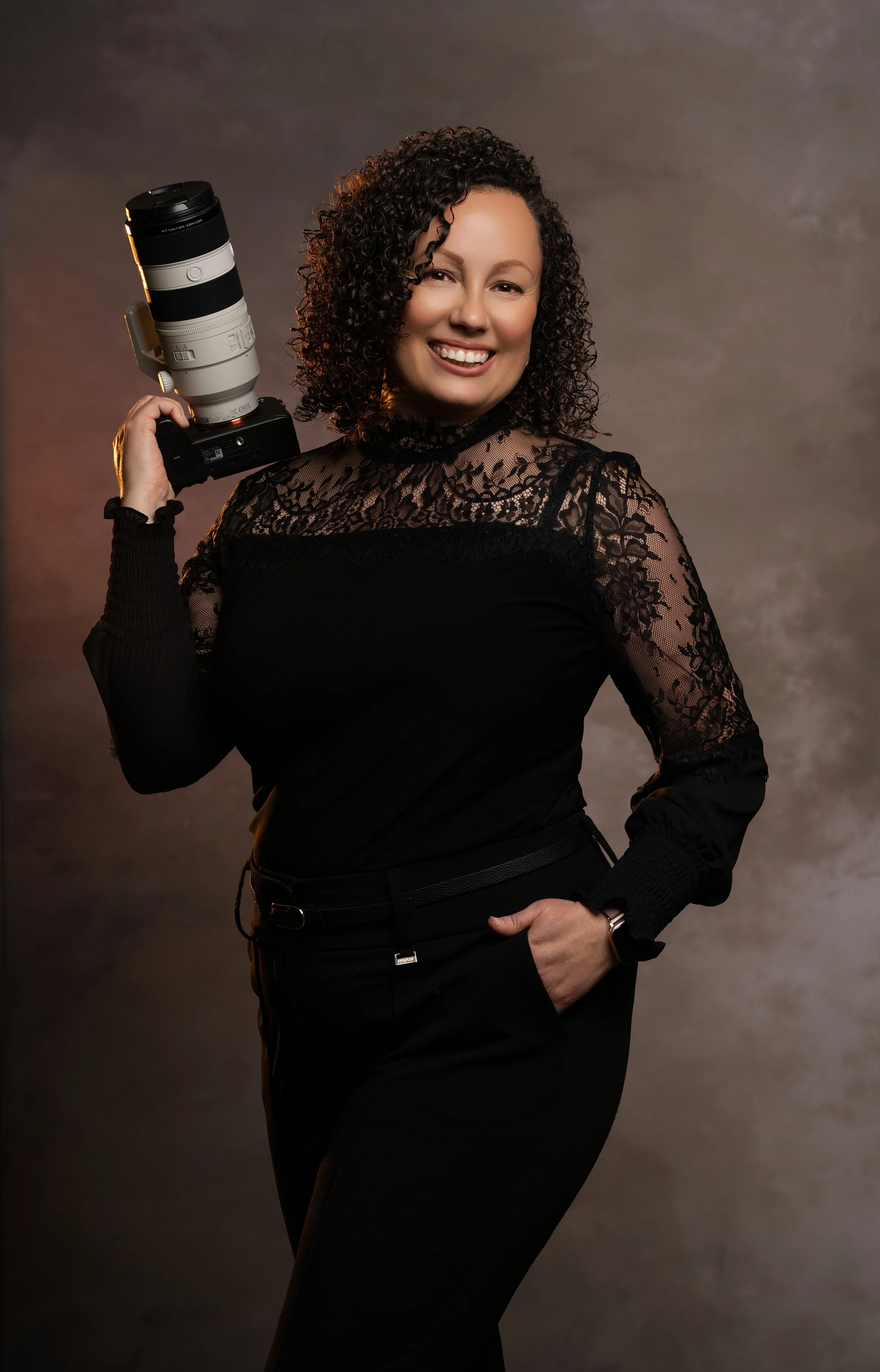 Woman with curly hair smiling, holding a large camera lens on her shoulder, wearing a black lace top and black pants, posing confidently in a studio setting.