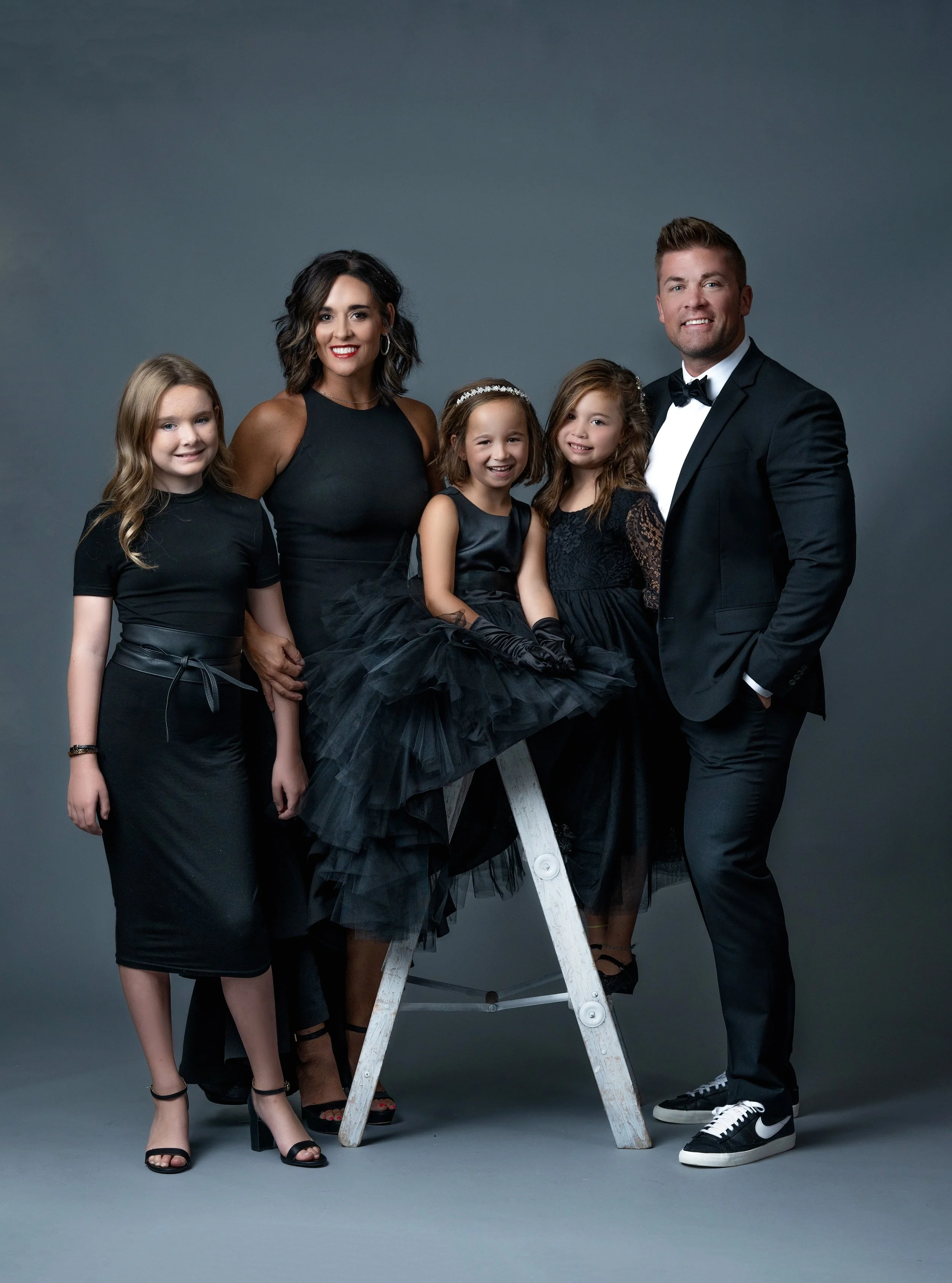 A group of five people dressed in formal black attire, standing around a stepladder against a plain gray background. They are smiling and looking at the camera. Omaha Nebraska family portrait photographer.