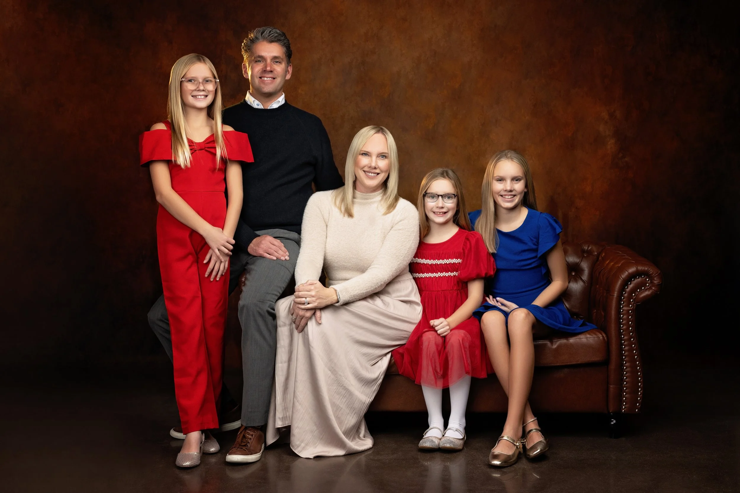 Family of five posing for a portrait against a dark brown background. The family includes a man, a woman, and three girls, all smiling. The man is wearing a black sweater over a white collared shirt, the woman is in a cream sweater and a long beige s