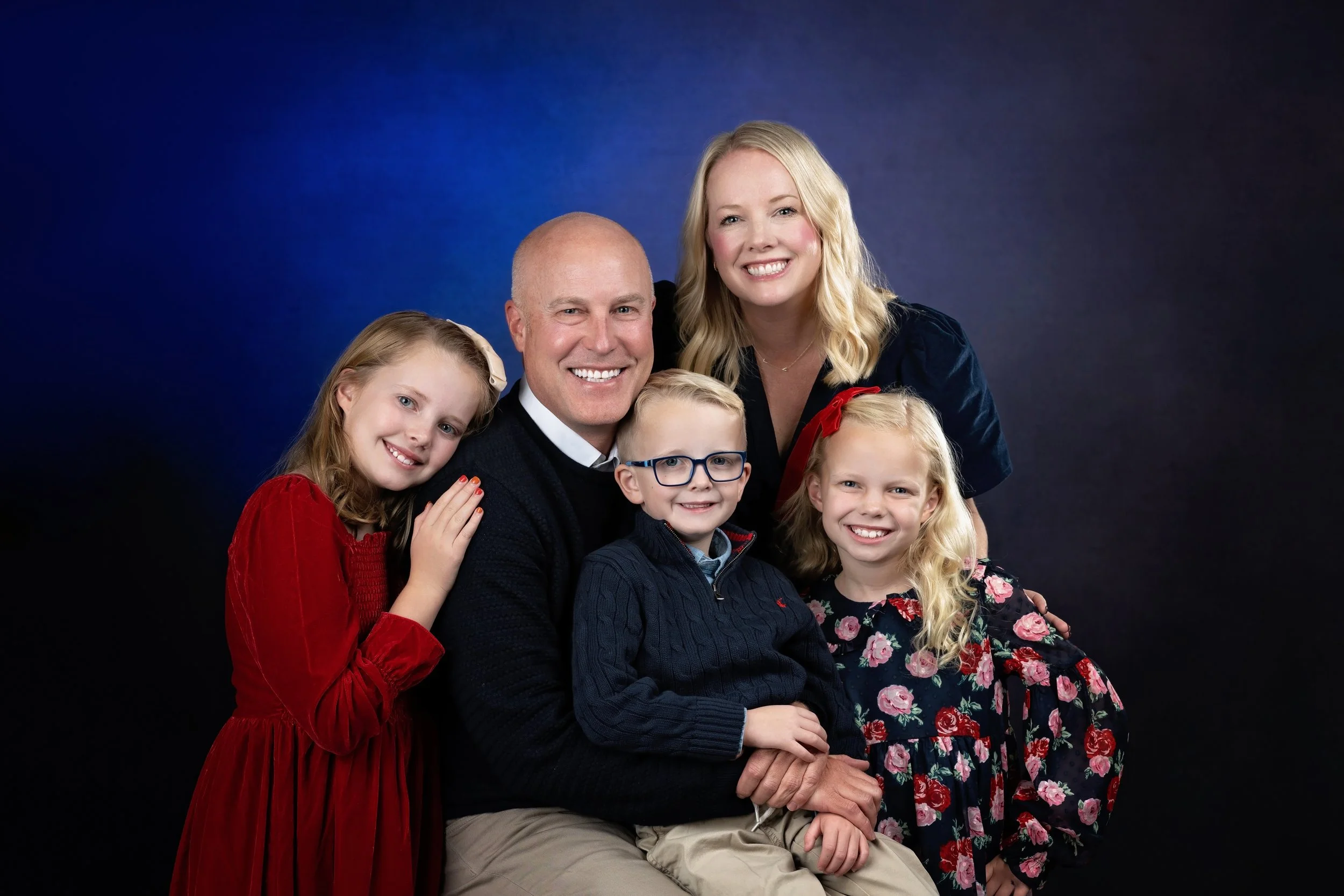 A family of six posing together against a dark blue background. The group includes a man, woman, and four children, all smiling and dressed nicely.