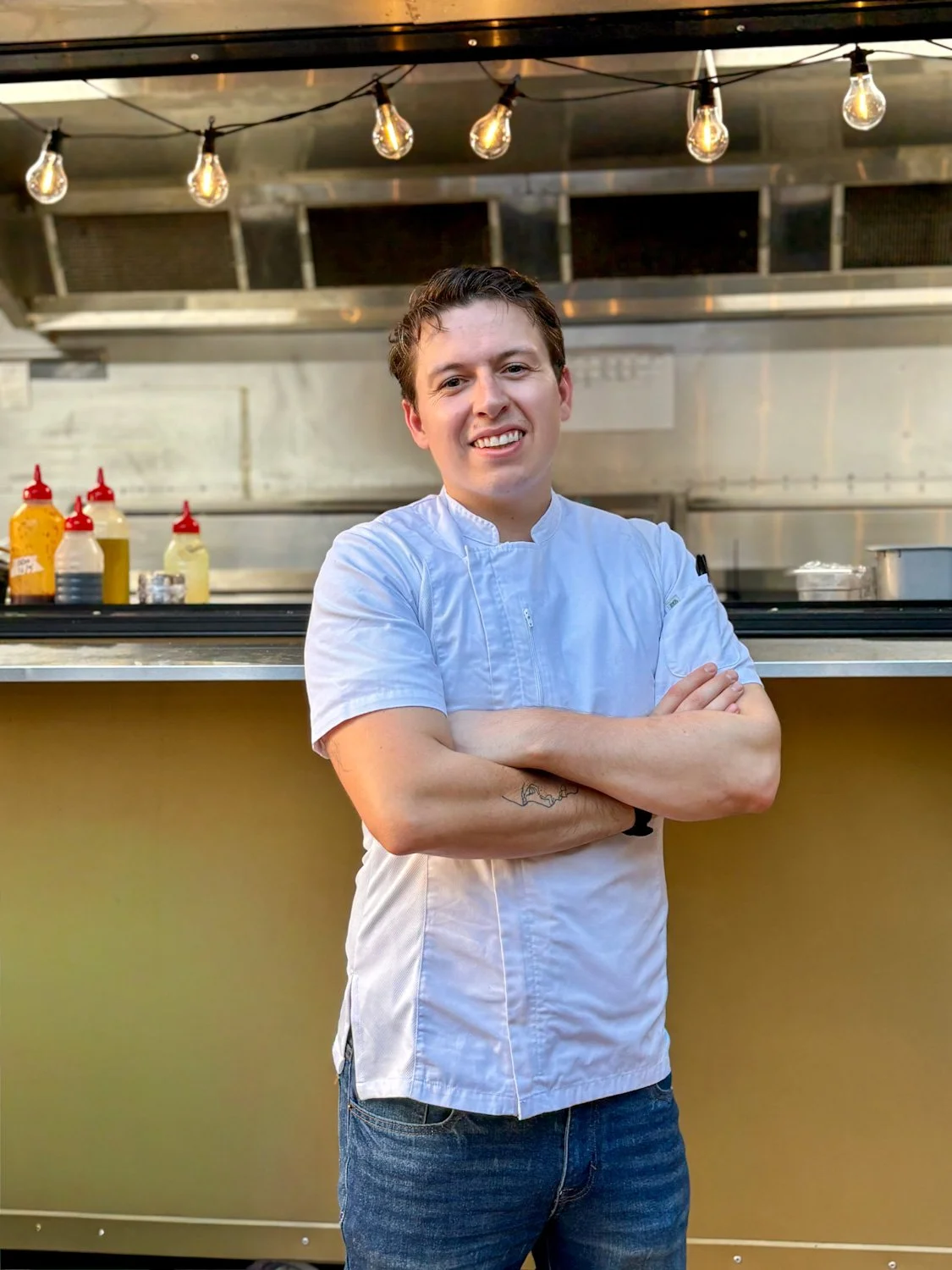A young male chef standing with arms crossed in a commercial kitchen, smiling. He is wearing a white chef's jacket and blue jeans, with ketchup and mustard bottles visible on the counter behind him.