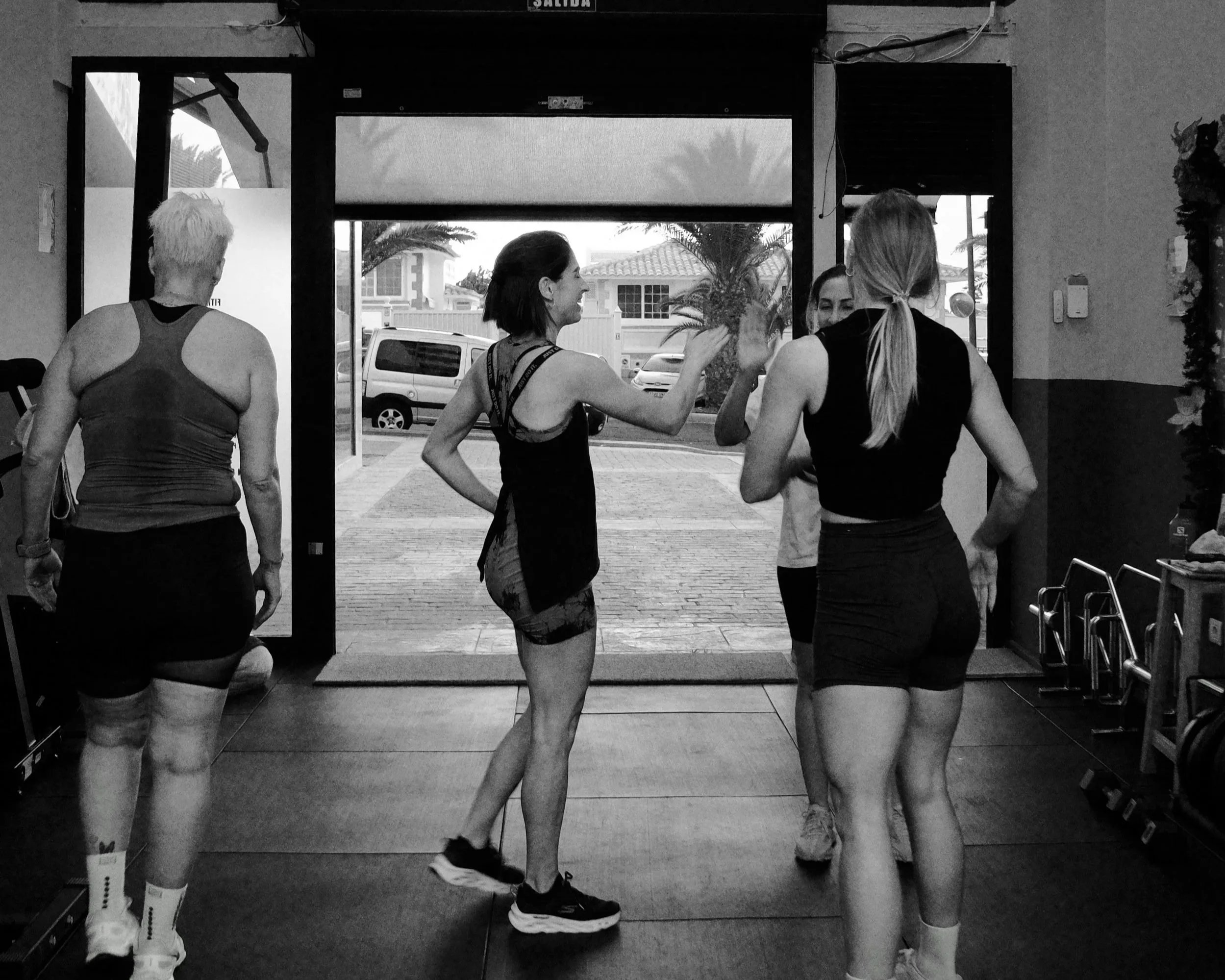 Four women in workout attire inside a gym, engaging in a high-five and conversation. Outside the gym, parked cars and palm trees are visible through the open door.