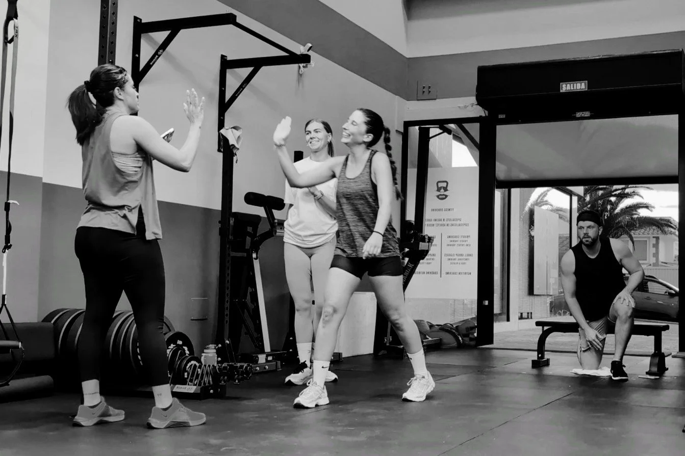 Four people in a gym giving high-fives. Two women in workout clothes stand in the center, one woman on the left, and a man on the right, sitting on a bench. Gym equipment is visible in the background.