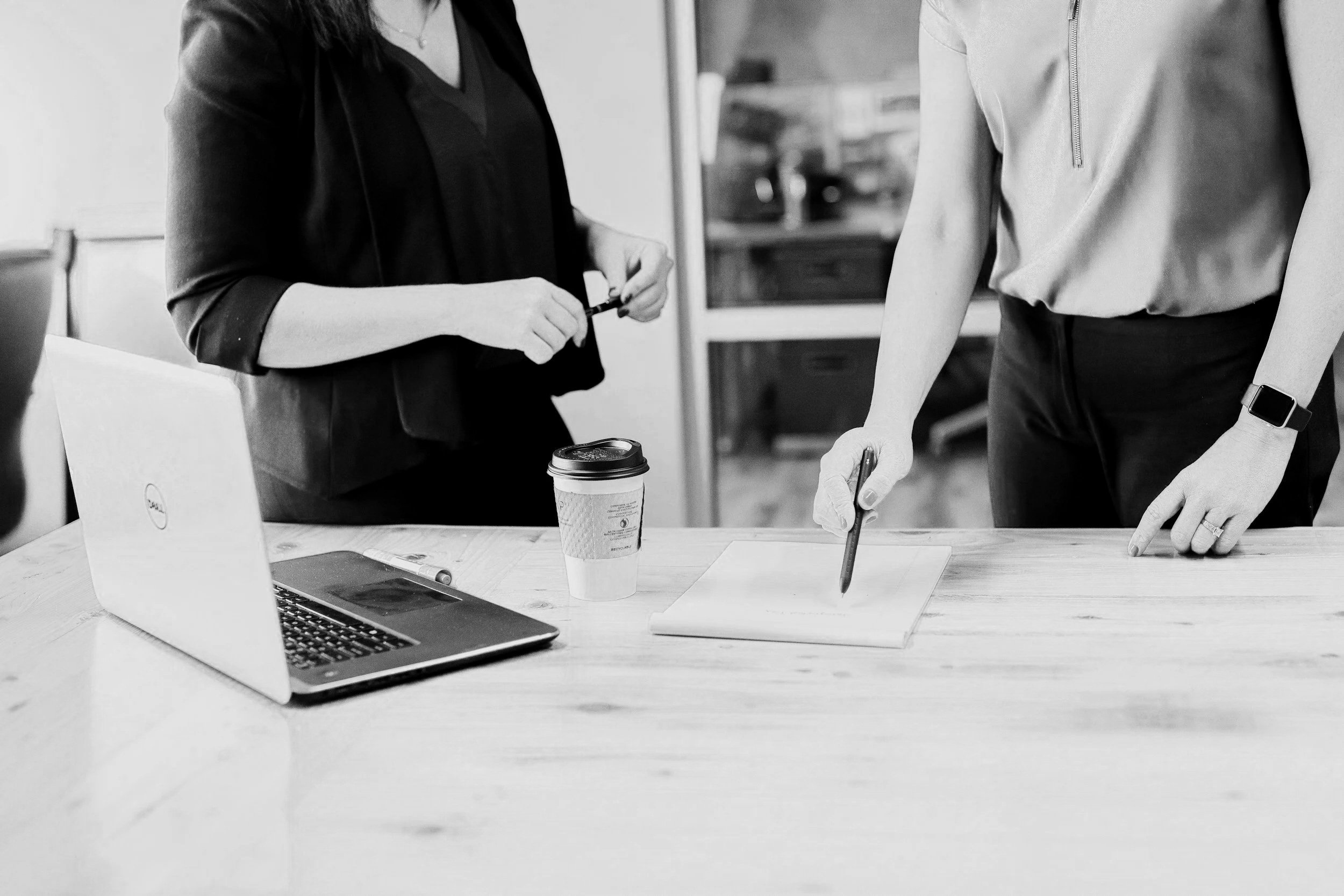 Two women in an office setting standing over a table, one appearing to be explaining or discussing something while the other writes on a notepad. On the table are a laptop, a pen, a disposable coffee cup, and a pen.