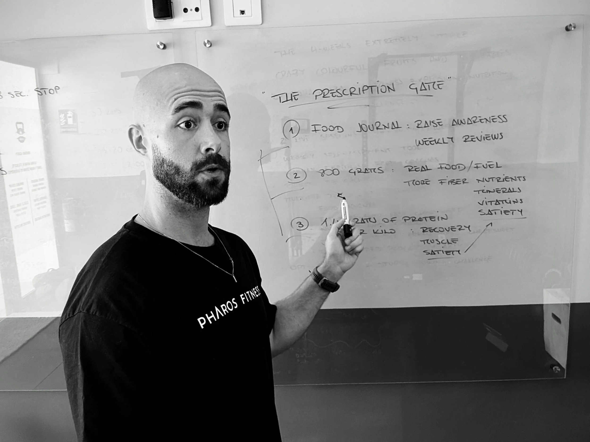 A man with a beard and bald head wearing a T-shirt that says 'PHAROS FITNESS' is standing in front of a glass whiteboard, holding a marker, and appears to be explaining or teaching. The whiteboard behind him contains handwritten notes and diagrams.
