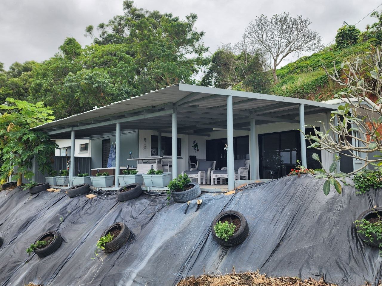 A house built on a hillside with a large covered porch, surrounded by lush trees, with tires and potted plants on a black tarp slope in the foreground.