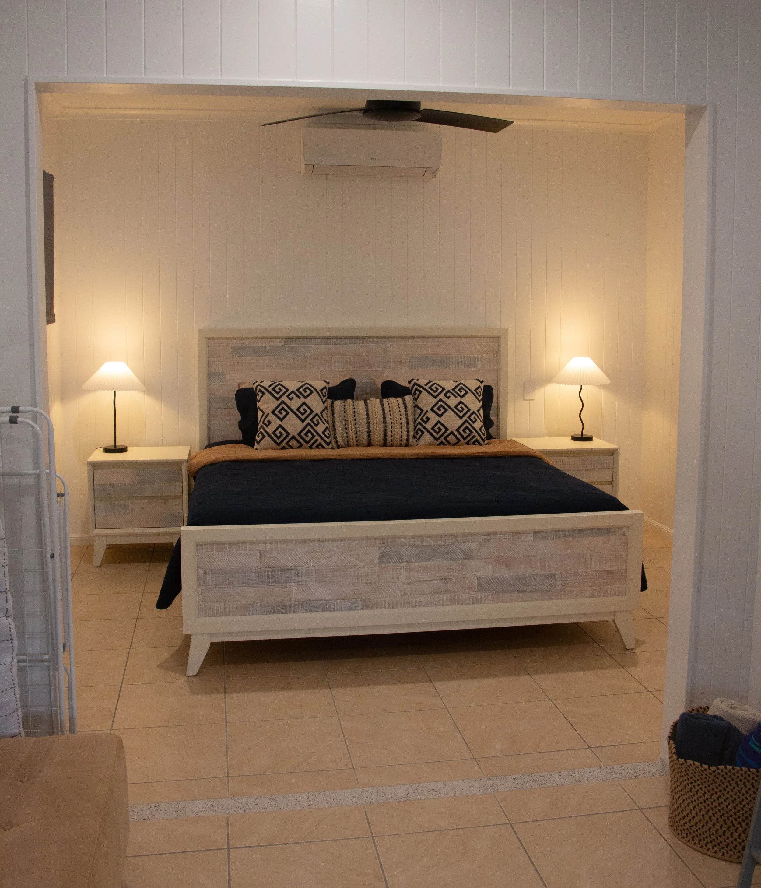 A bedroom with a bed, pillows, two lamps, a nightstand, a ceiling fan, and an air conditioning unit.