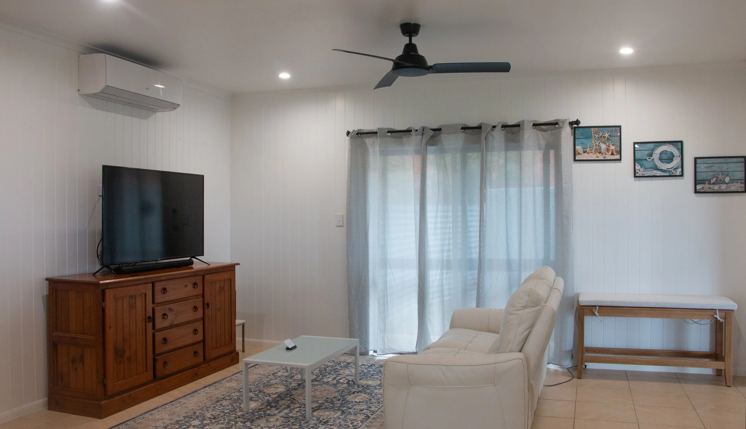 Living room with white walls, a black ceiling fan, a flat-screen TV on a wooden cabinet, a white couch, a small white coffee table, and curtain-covered window with seaside-themed framed pictures on the wall.