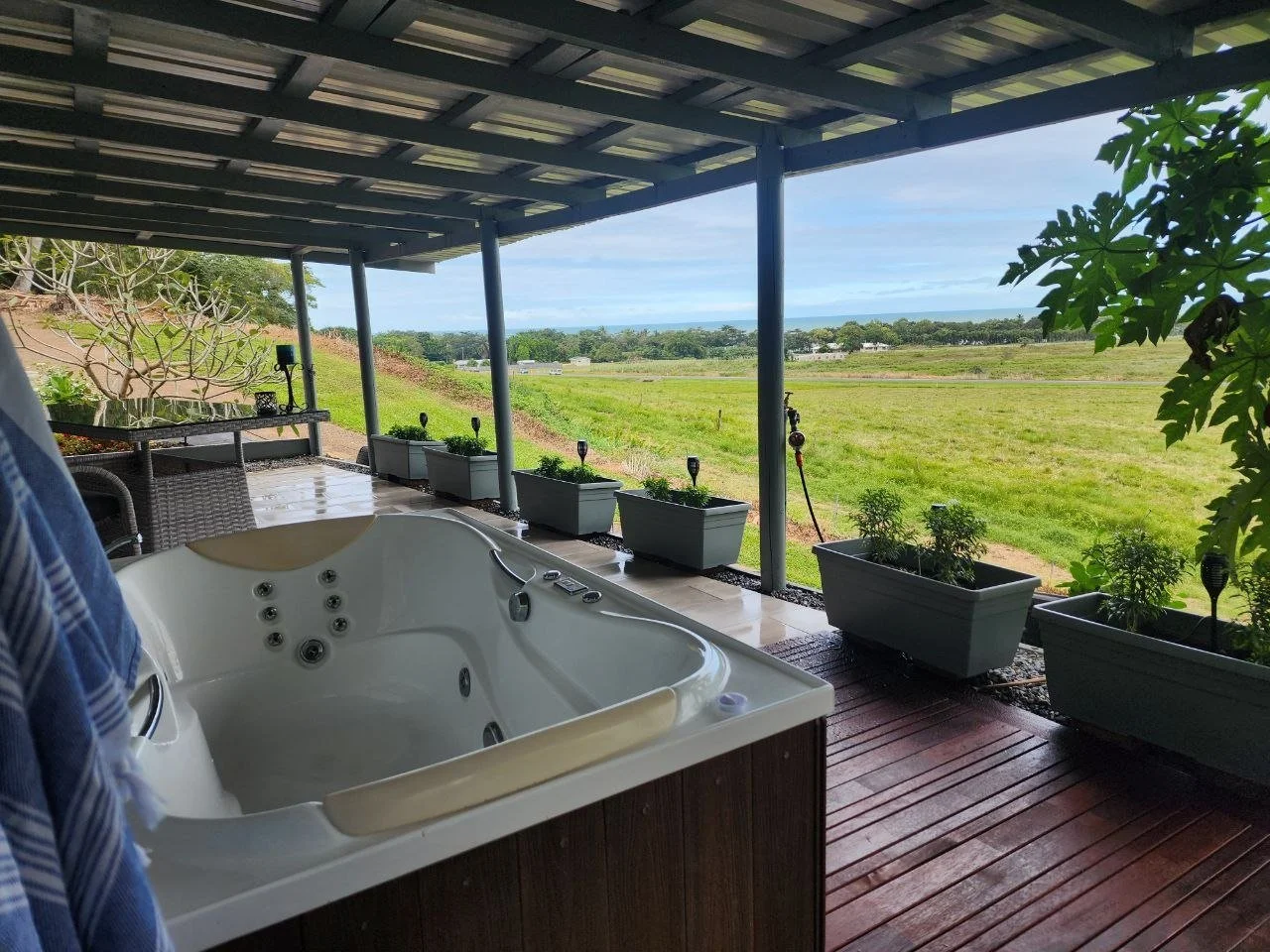 Outdoor patio with hot tub and potted plants overlooking a grassy field and distant trees under a covered roof.