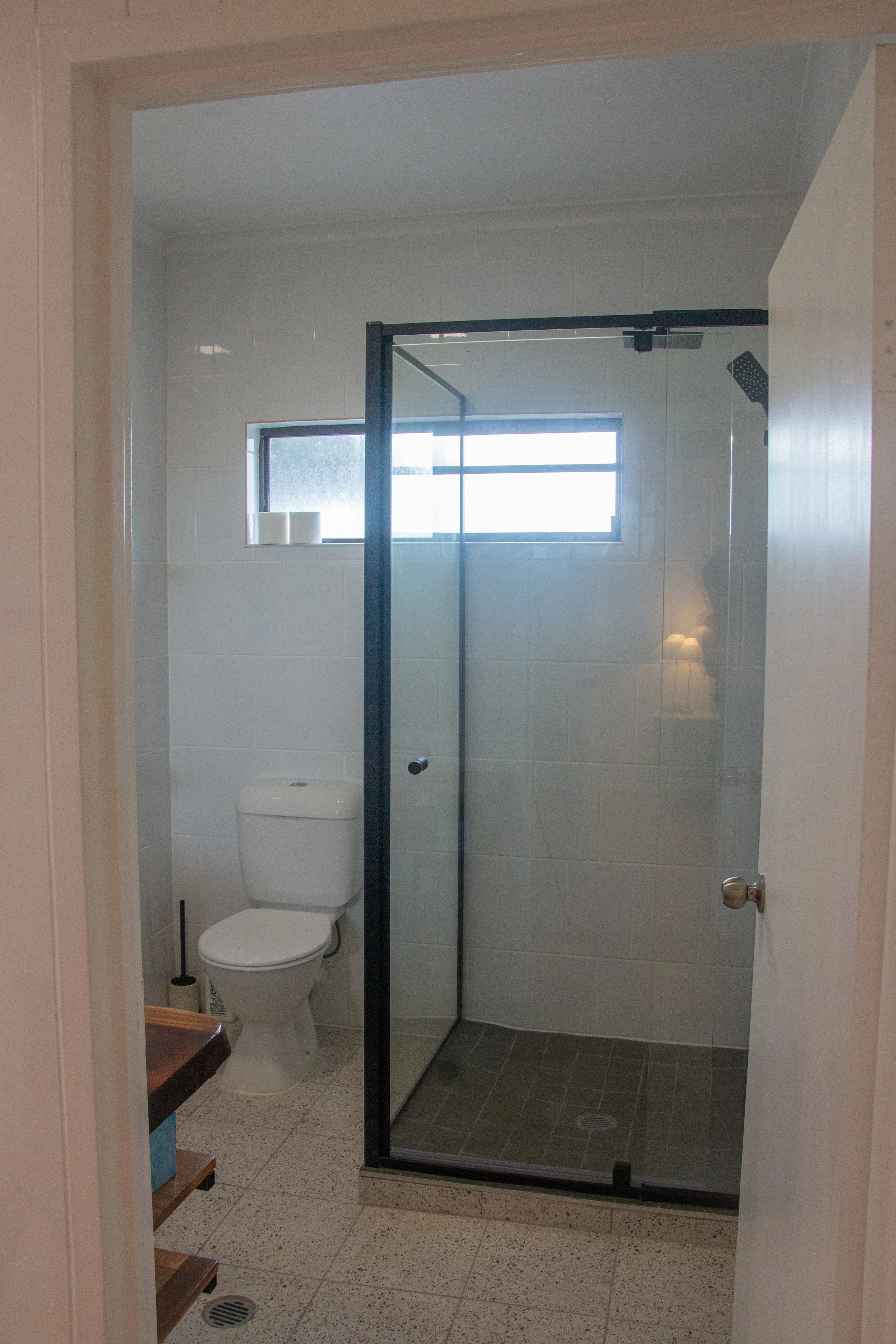 A small bathroom showing a white toilet, a glass shower enclosure with a black frame, a window with frosted glass, and a tiled floor.