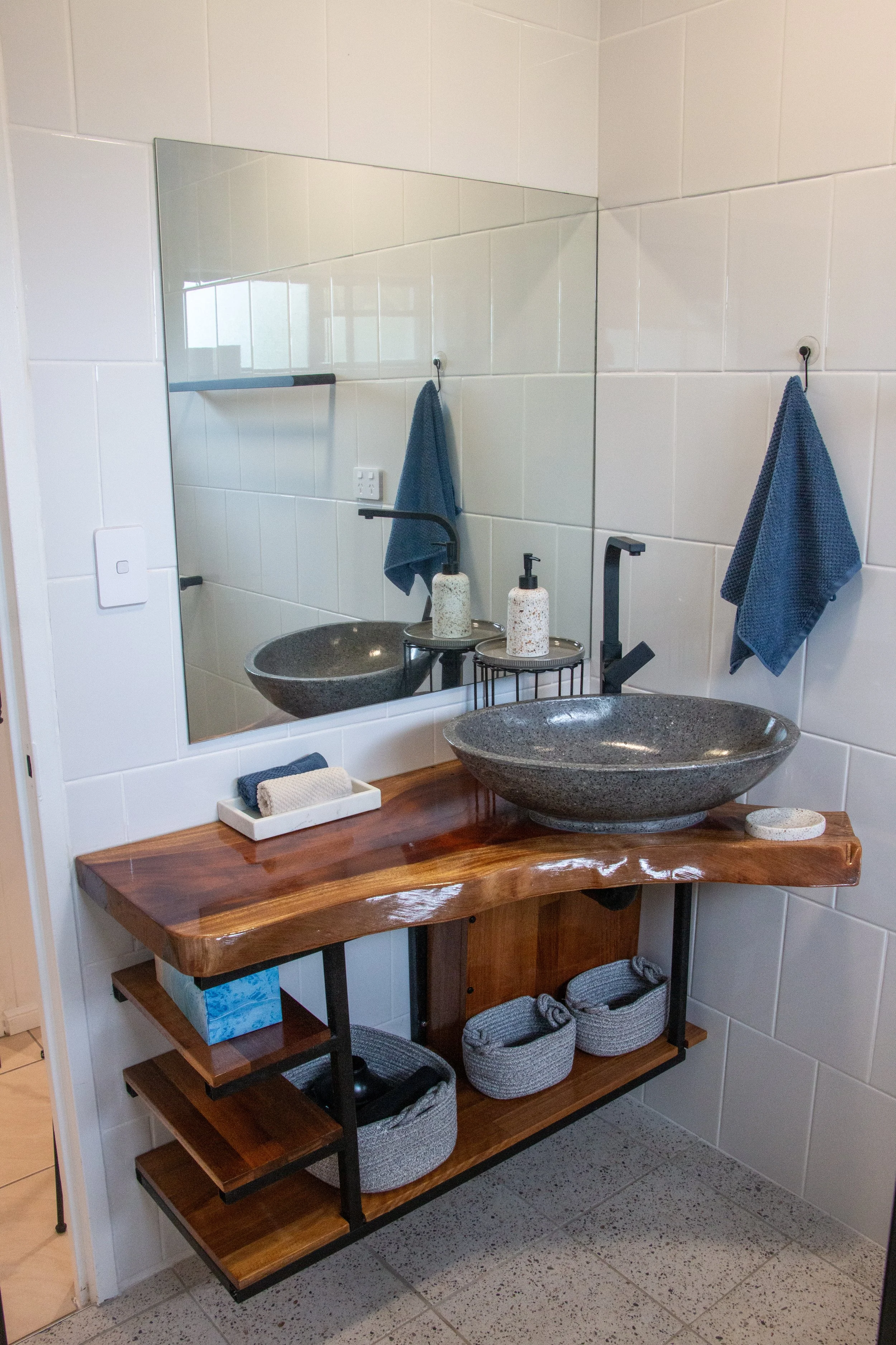 A bathroom vanity with a wooden countertop, a black vessel sink, and a mirror above it. Two blue towels are hanging on hooks on the white tiled wall. There are soap dispensers on the countertop, one with black and one with white and speckled patterns. Below the wooden countertop, there are open shelves holding baskets, boxes, and other items.