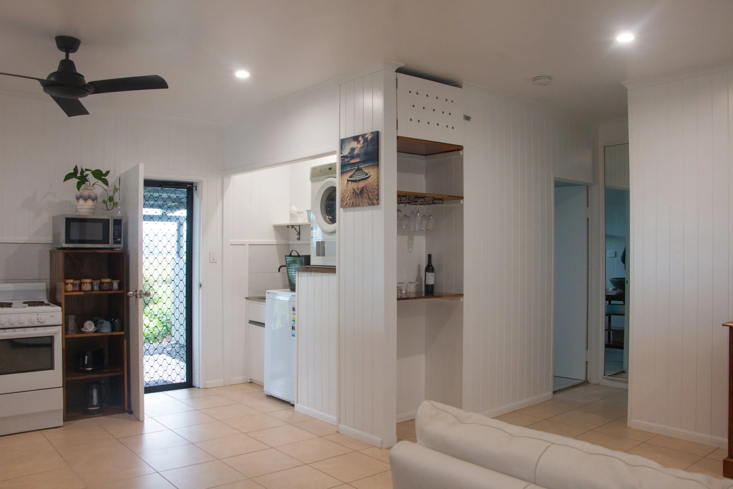 A kitchen and living area with white paneled walls, beige tiled floor, a ceiling fan, and built-in open shelves. Visible appliances include a microwave on a wooden stand, a stove, a washing machine, and a mini-fridge. There is a door with a security screen leading outside, and a decorative beach scene painting on one wall.