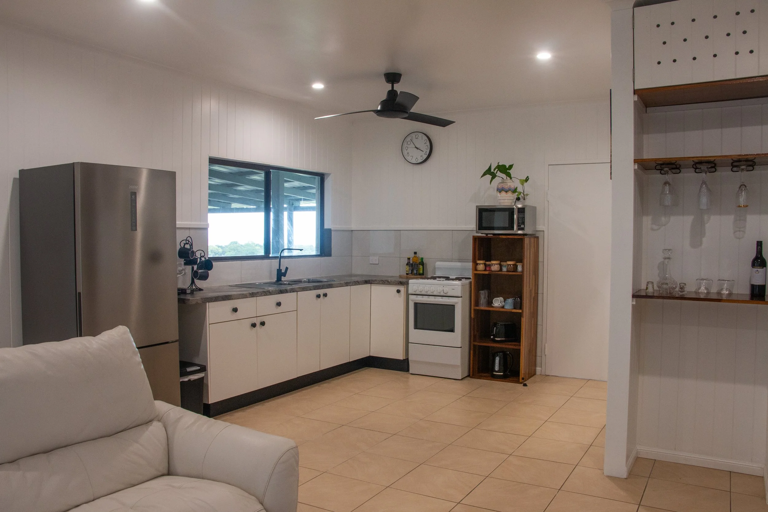 View of a modern kitchen with white cabinets, a stainless steel refrigerator, a microwave on a wooden shelf, a white stove, a window, and a black ceiling fan. In the foreground, part of a white sofa is visible.