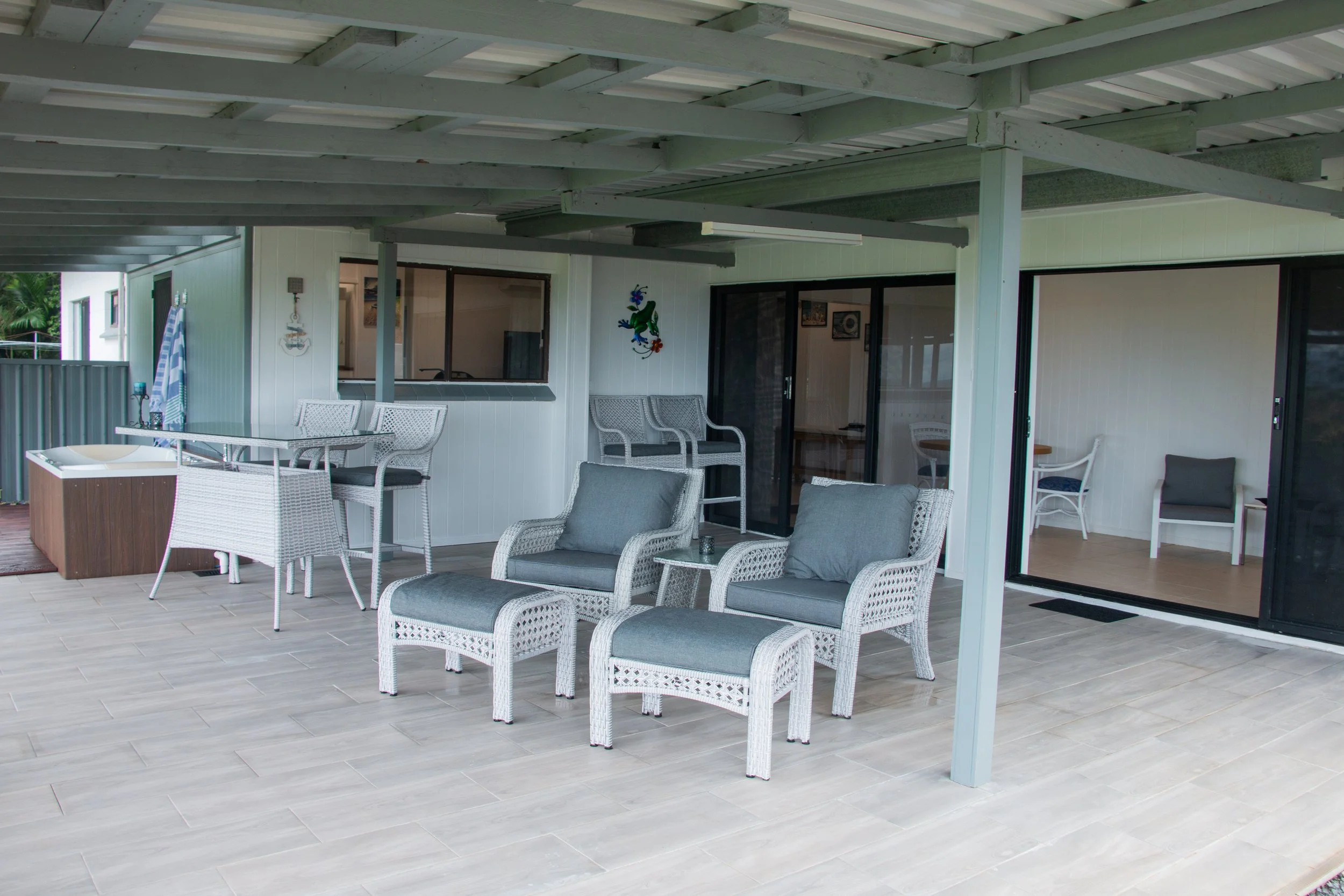 Covered outdoor patio area with wicker chairs, a table with chairs, and a hot tub, attached to a house with sliding glass doors and windows, in a tropical setting.