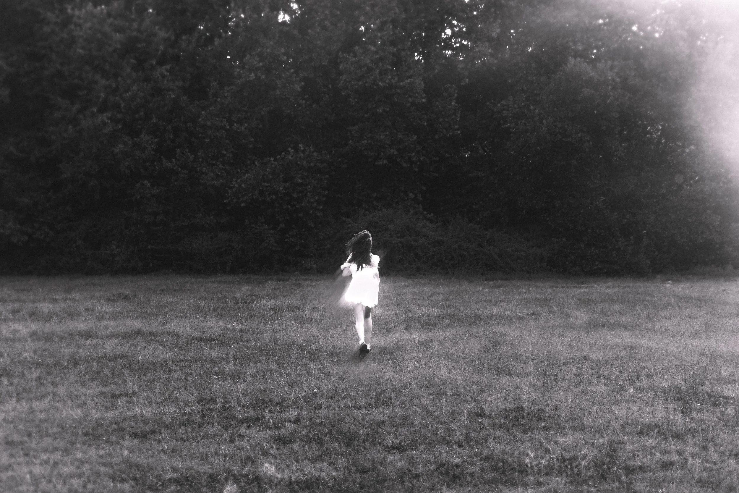 Una niña de espaldas corriendo en un campo con árboles al fondo, en una imagen en blanco y negro.