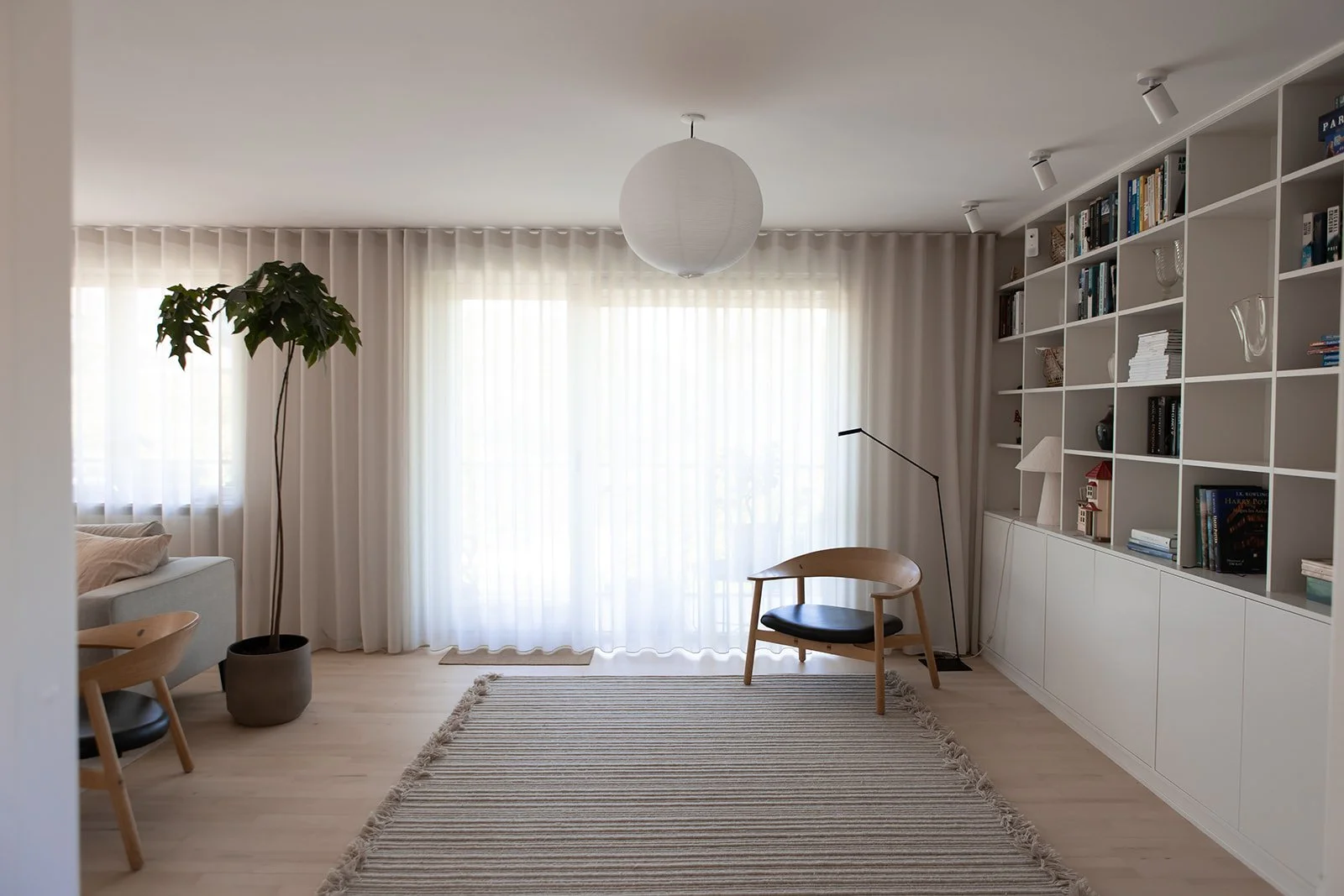 Stue med store vinduer med hvide gardiner, en pæreformet loftlampe, en stor grøn plante i potte, en beige sofa med puder, et sofabord, en væglampe, bøgernes reol, en lænestol, en stribet tæppe på lyst gulv.