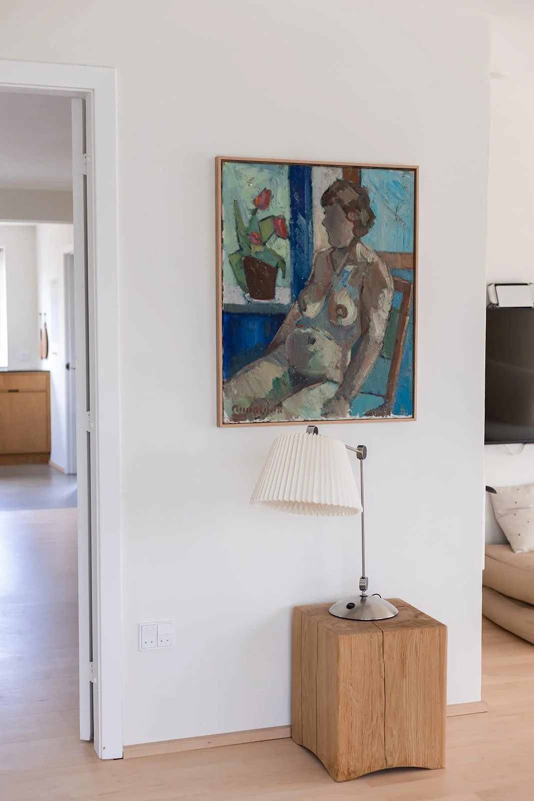 Et maleri af en nøgen person hænger på en hvid væg over en lille træbord med en moderne bordlampe. Der er et skab og en sofa i baggrunden.