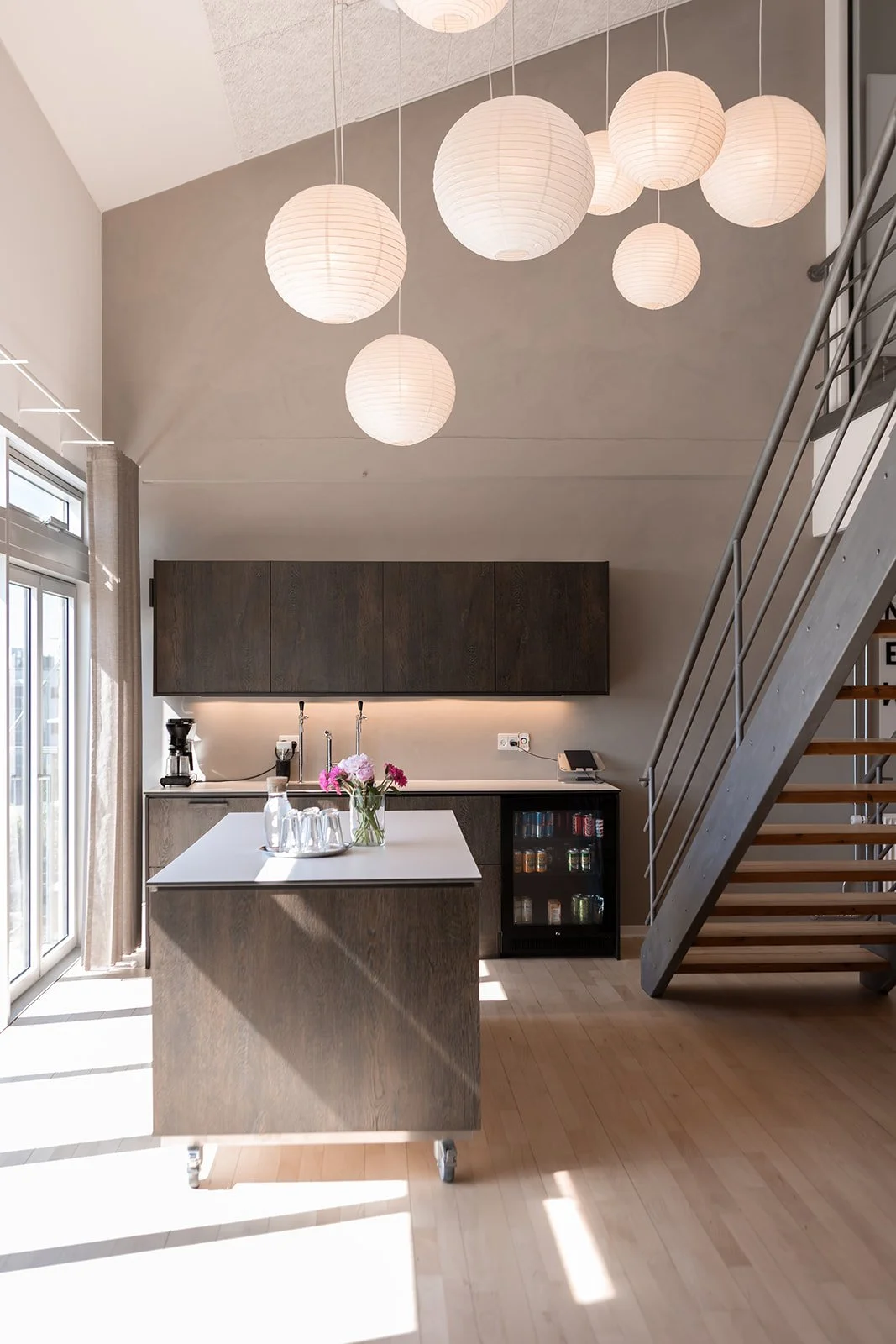 Moderne køkken med hængende luminaires og trappeløb i metal, køleskab med drikkevarer, og bord med blomster.