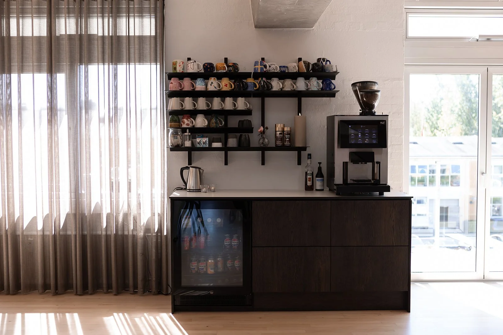 Kaffeestation med kaffeautomat, genstande på hylder, køleskab med sodavand, og vinduer med gardiner i baggrunden.