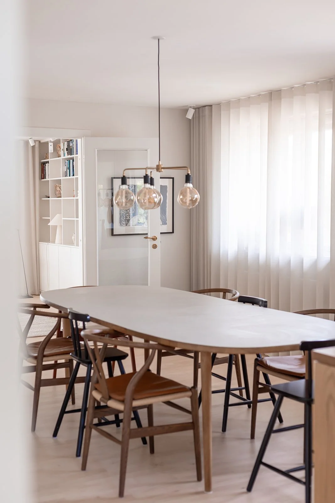 Samlet spisebord med otte forskellige stole i et lyst og moderne rum med store vinduer og gardiner.