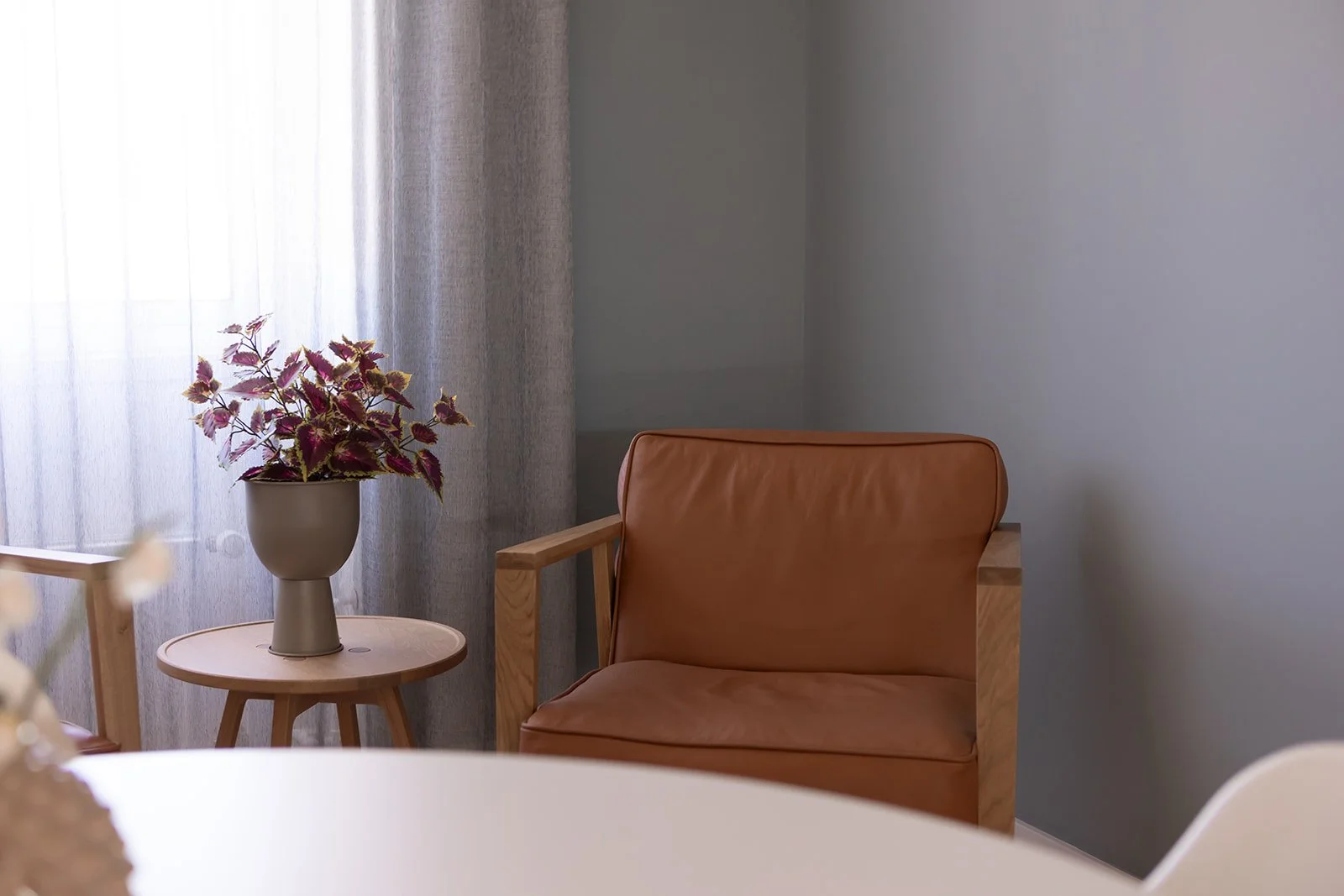 Et hjørne med en brunt lænestol, en rund træbord med en grå vase med lilla og grønne blomster, lette gardiner og en grå væg.