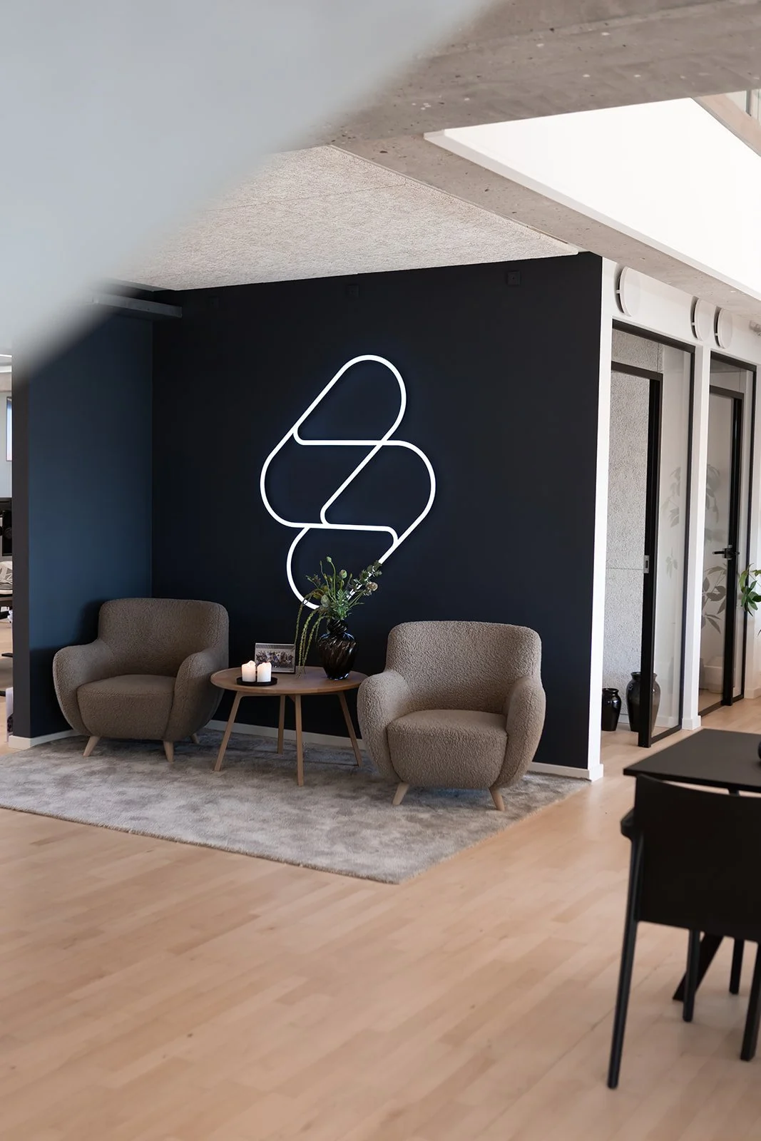 Et hyggeligt venteområde eller lounge med to beige komfortable armstole, et rundt træbord med stearinlys og en vase med grønne planter, frontet af en mørk væg med en moderne hvid neon-lysdekor i form af løse kurver.