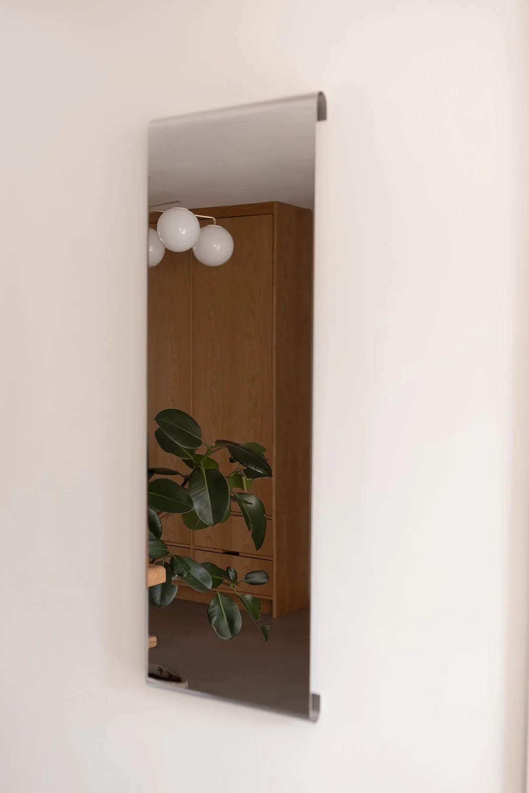 Refleksion i en vægmonteret spejl, der viser et rum med en grøn plante, et brunt skab og en loftlampe med hvide globusformede pærer.