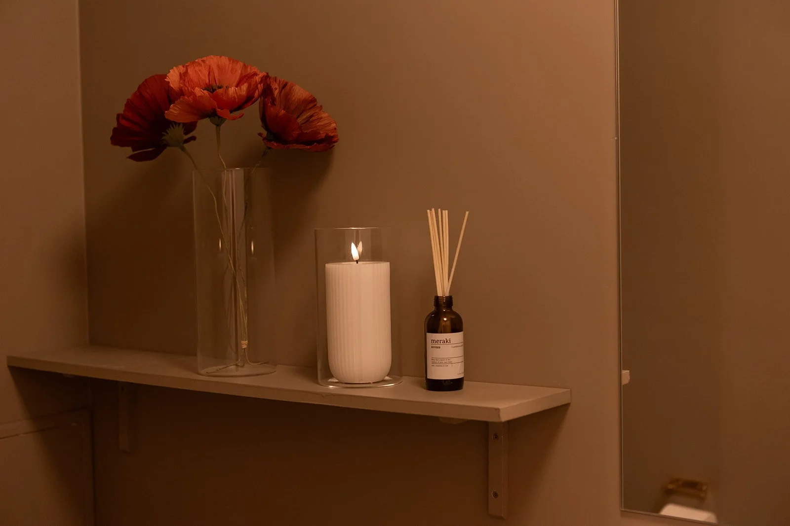 Vase med orange-poppede blomster, lysende stearinlys i glasholder, en med duftdiffuser i en lille brun flaske, placeret på en hylde mod en beige væg.