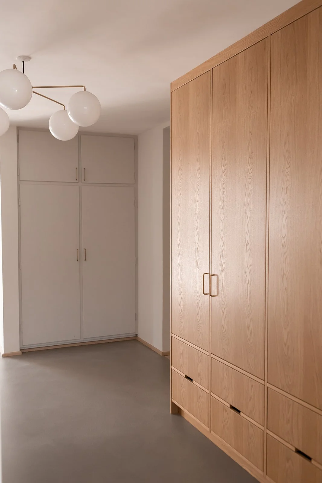 Et hjørne af et værelse med en stor træskab og en hvid væg. Der er en loftlampe med globus-armatur. Gulvet er gråt.