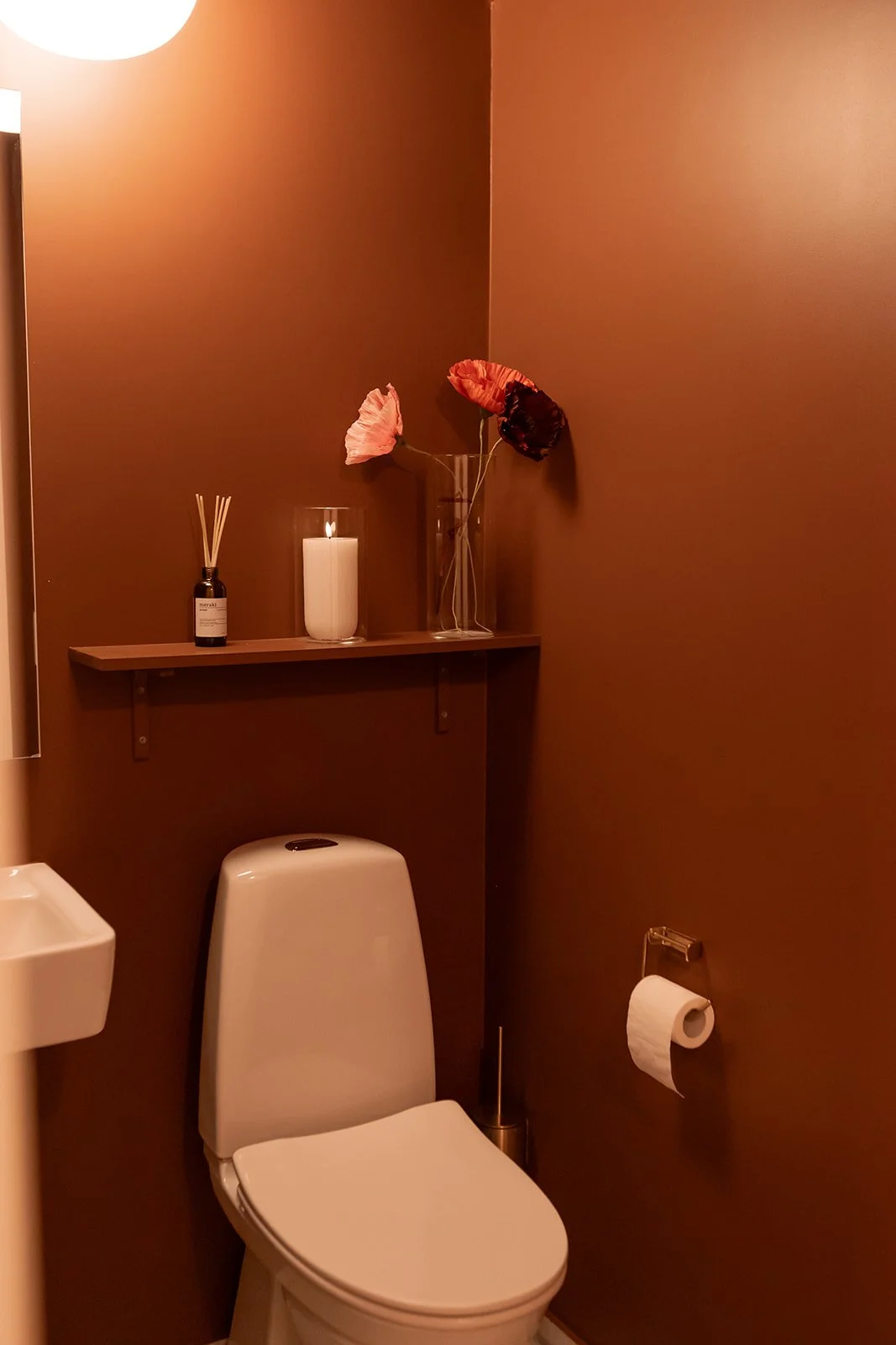 Badeværelse med brun væg, toilet, toiletpapirholder, lysende stearinlys, vase med kunstige blomster, diffuser med dufte. Skum affald på toilettet.
