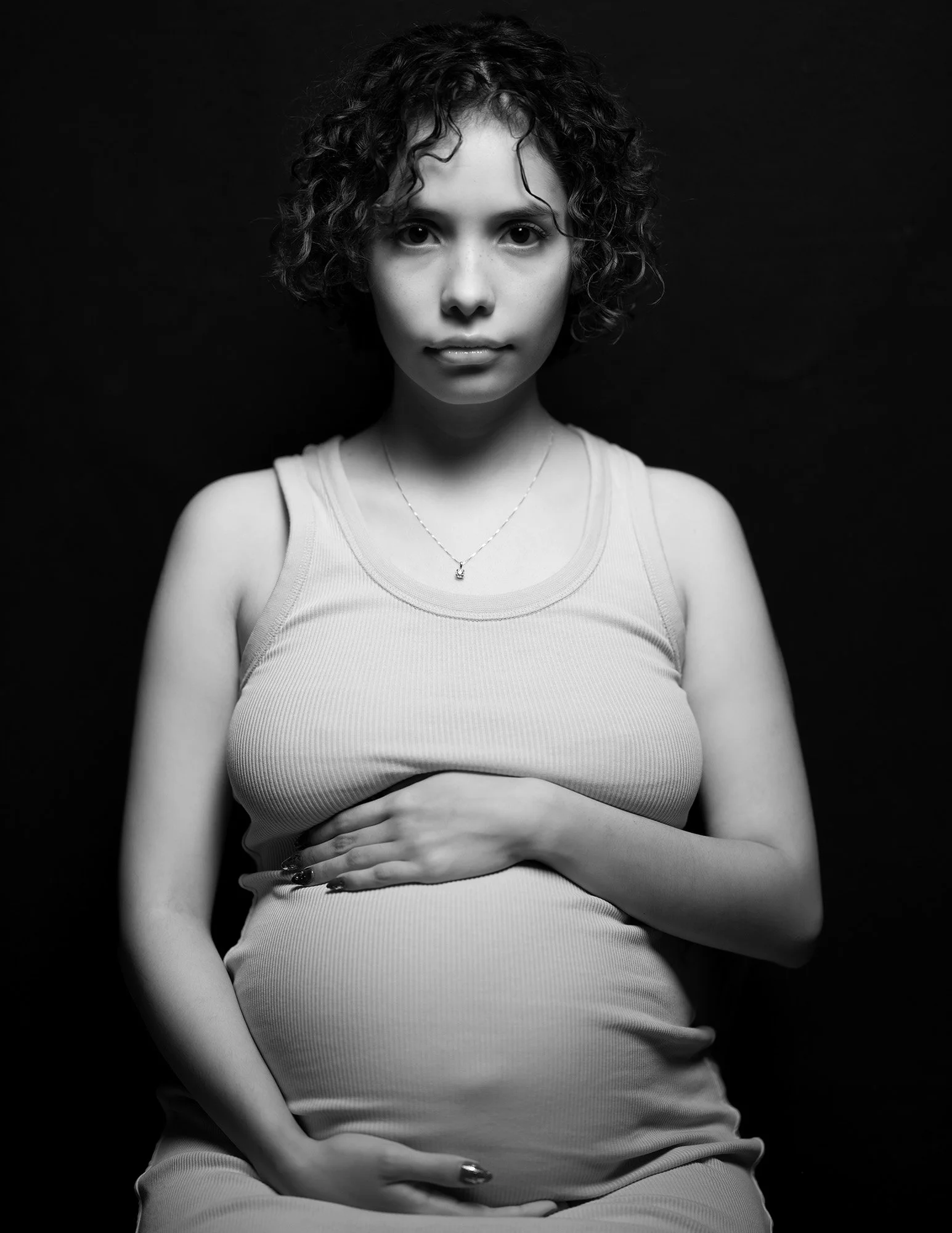 Black and white portrait of a pregnant woman with curly hair wearing a tank top, with one hand resting on her belly and the other on her chest against a dark background.