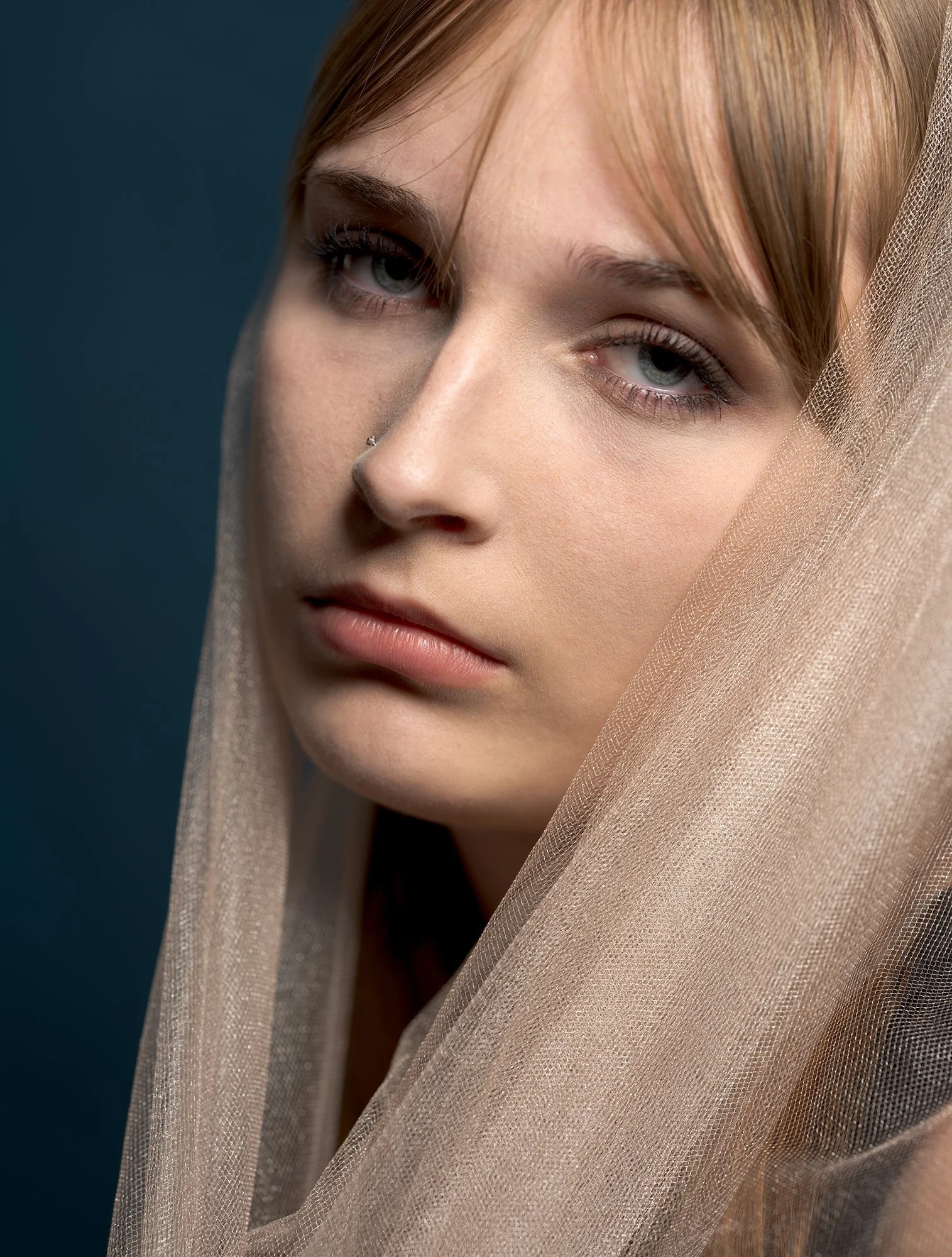 Close-up portrait of a young woman with light skin, blue eyes, and light brown hair, with a sheer beige fabric draped around her head.