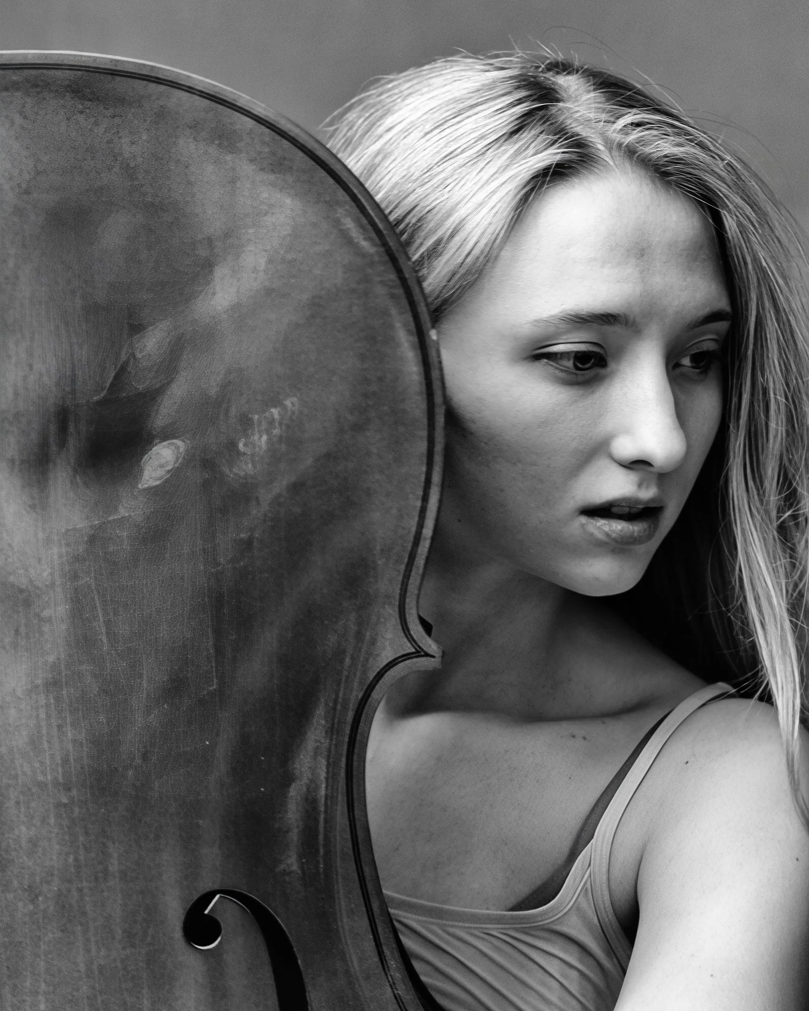 Black and white photo of a woman with blonde hair and a serious expression, partially obscured by a large musical instrument, likely a cello.
