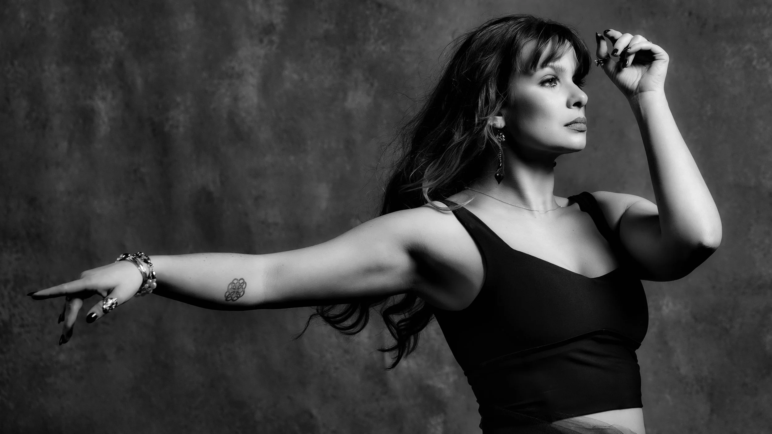 A black and white photo of a woman with wavy hair and jewelry, extending her arm and looking to the side against a textured background.