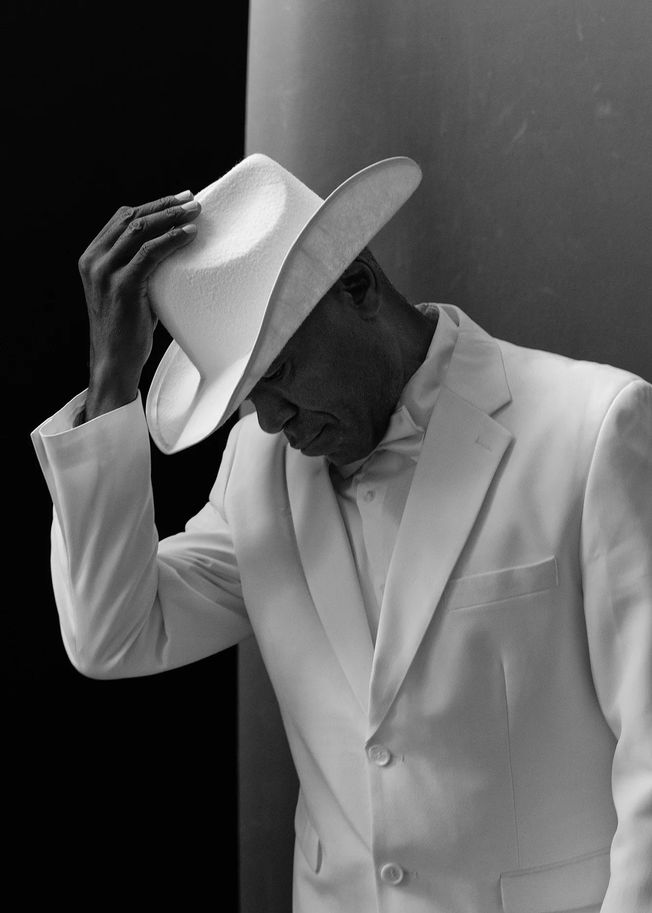 Black and white photo of a man wearing a white suit and a large white hat, tipping the hat downward, with a contemplative expression.