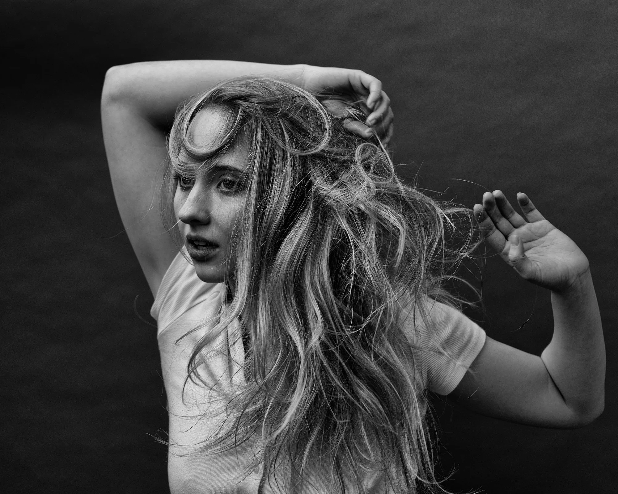 Black and white photo of a woman with long, wavy hair, posing with one arm over her head and the other hand near her face, against a dark background.