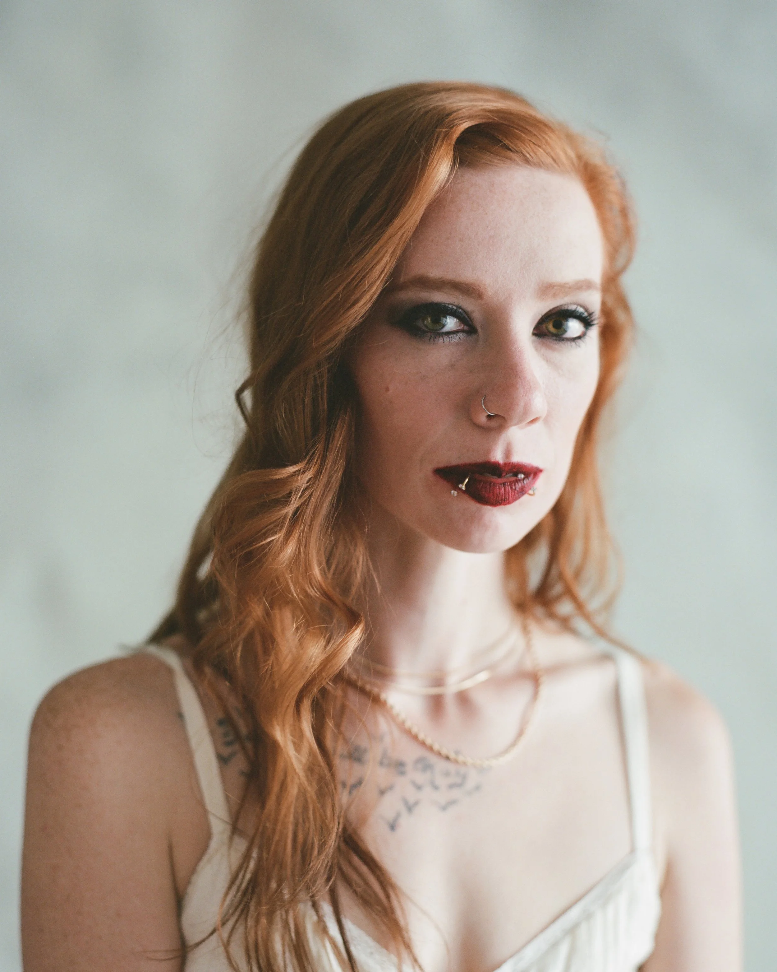 A young woman with red hair, green eyes, tattoos on her shoulder, wearing makeup, gold necklaces, a nose ring, and dark lipstick.