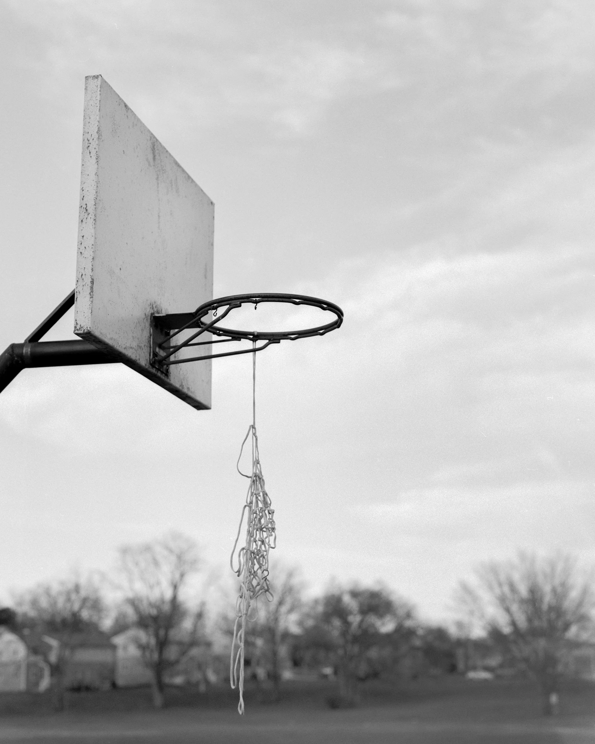 A basketball hoop with a backboard and net, hanging outdoors against a cloudy sky with trees and a road in the background, in black and white.