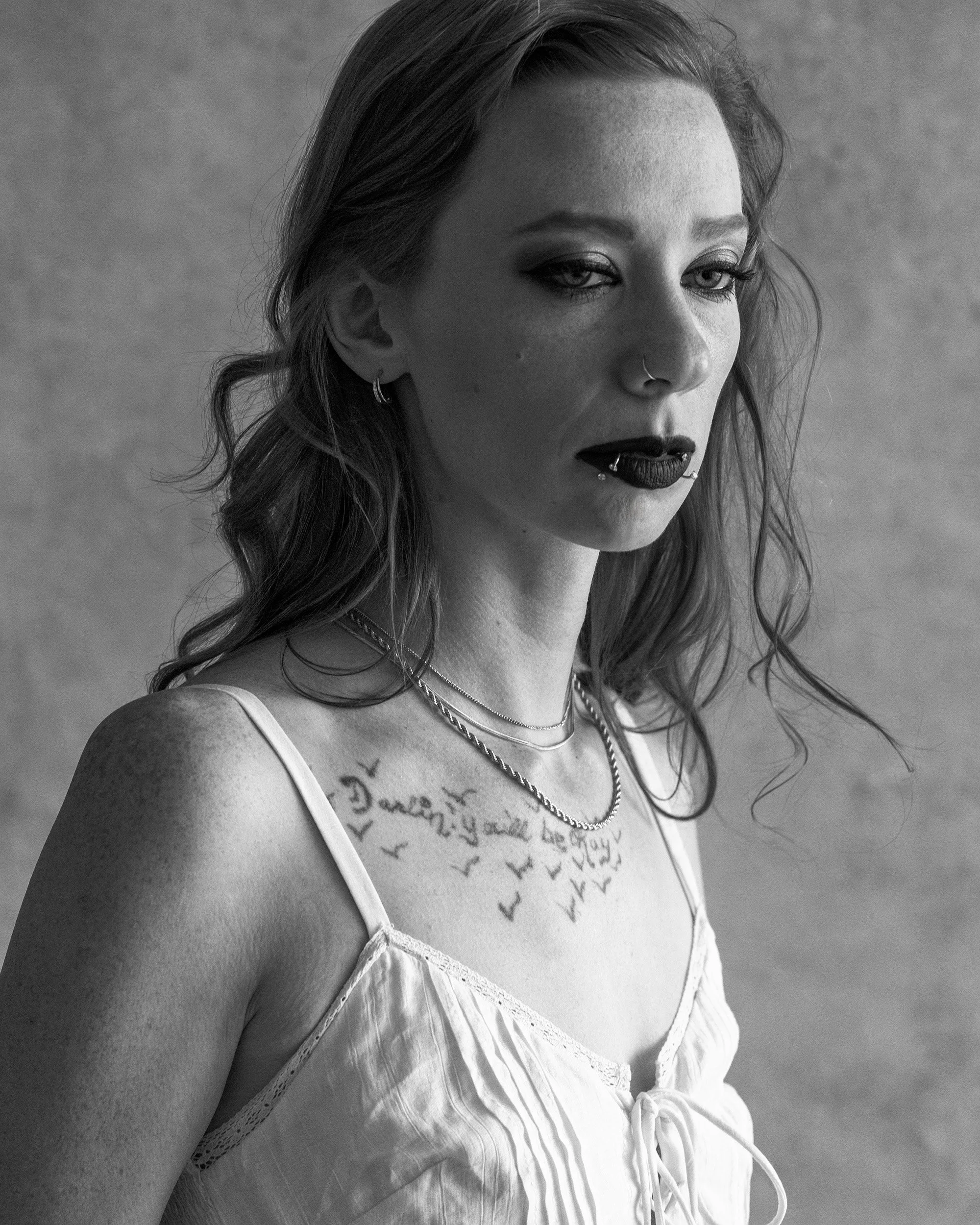A black and white portrait of a woman with long, wavy hair, wearing a white lace-trimmed dress, layered necklaces, and multiple facial piercings, with text tattoos on her chest.