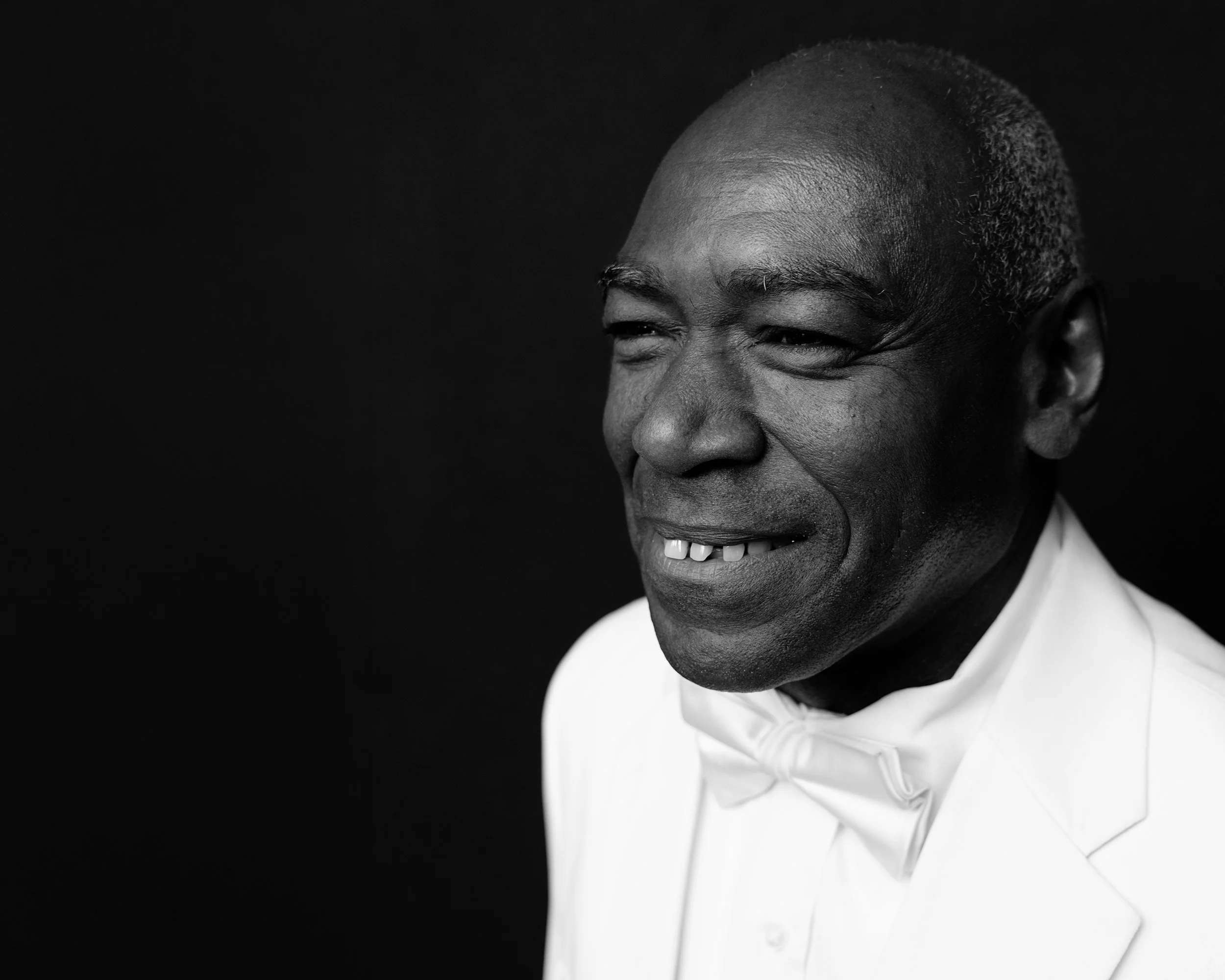 Close-up black and white photograph of a smiling man with short, graying hair wearing a white collared shirt.