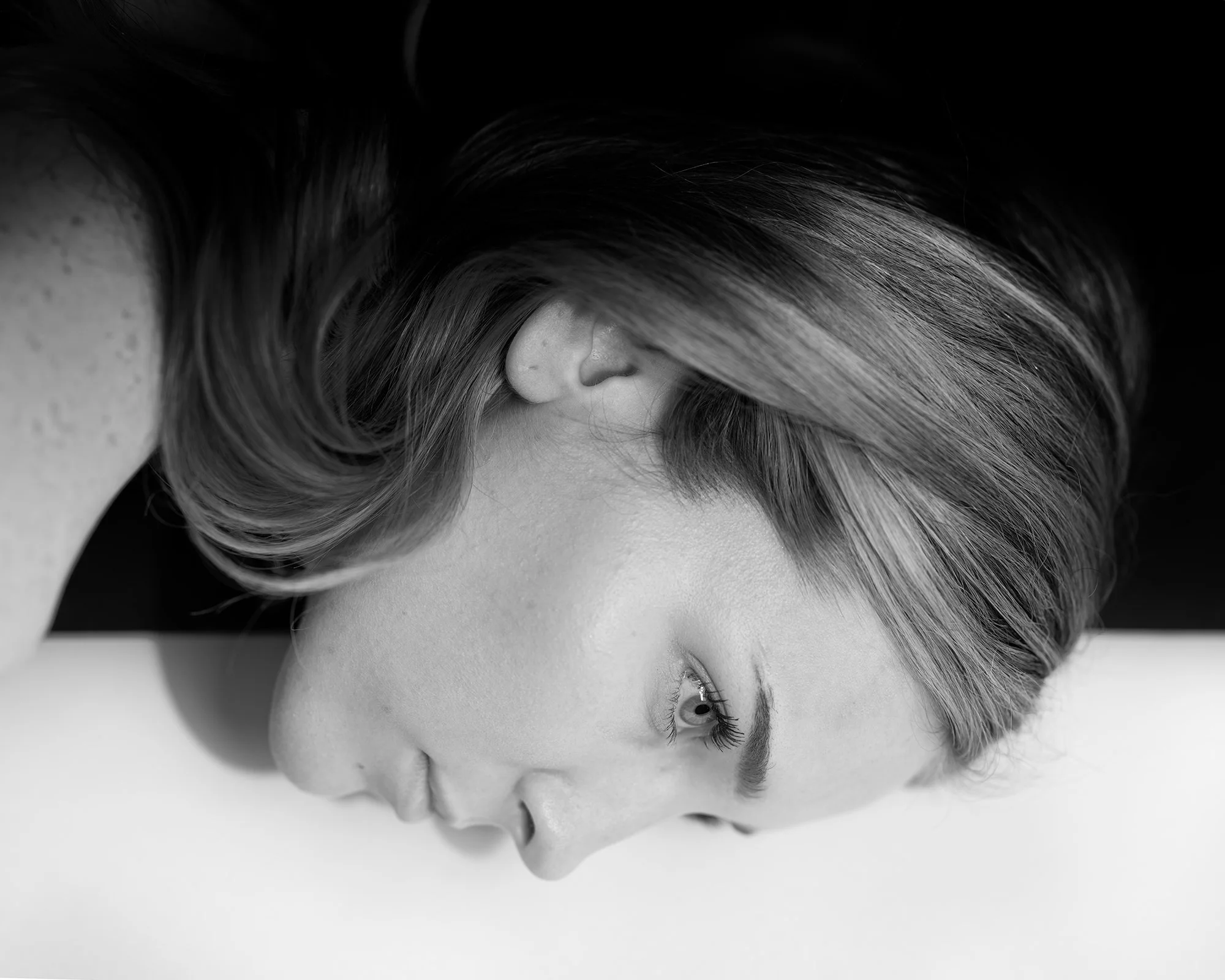 A woman with light skin and straight hair lying on a surface, with her head turned sideways, in black and white photography.