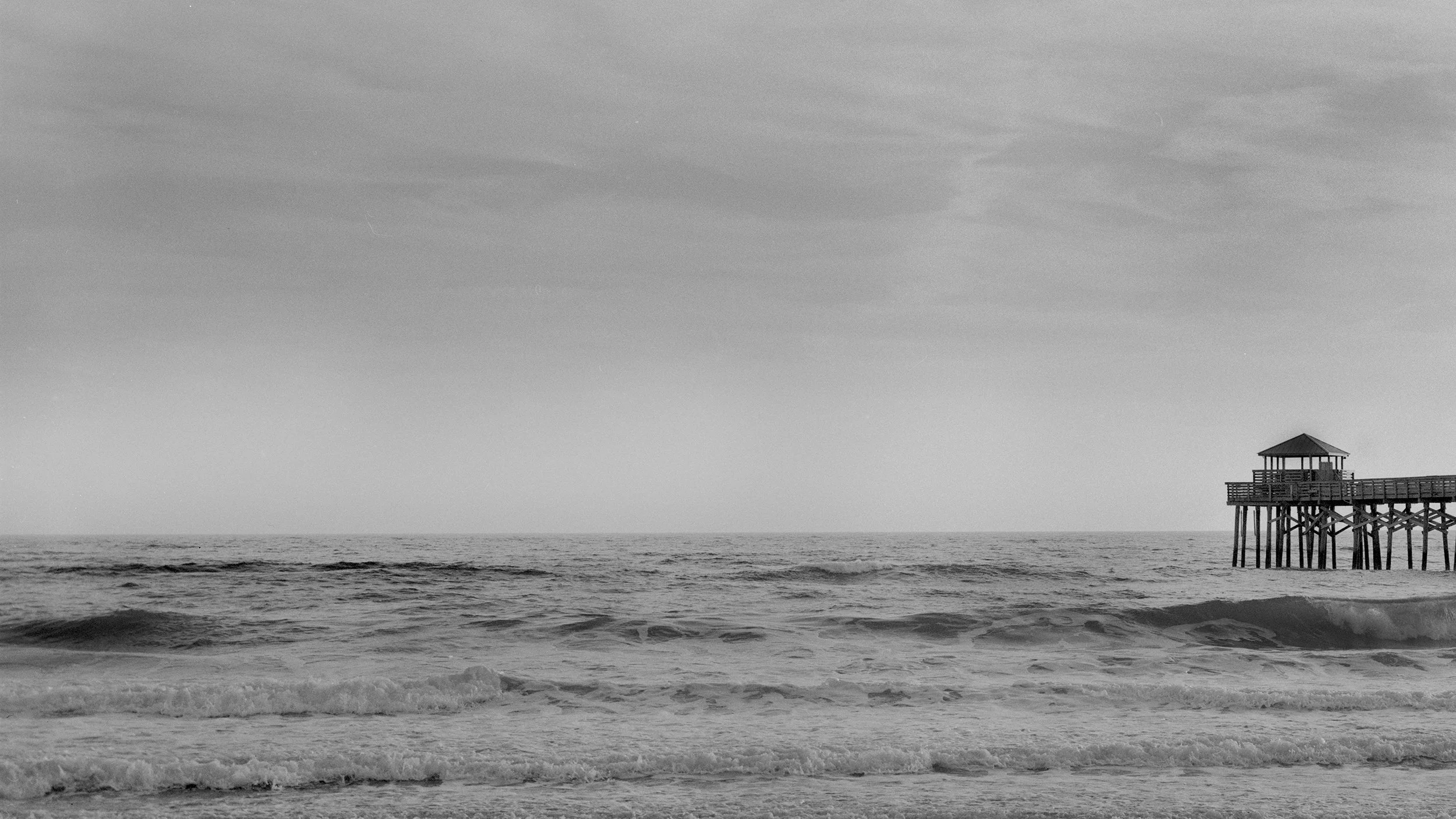 A black and white photo of an ocean with waves and a wooden pier with a small gazebo at the end.