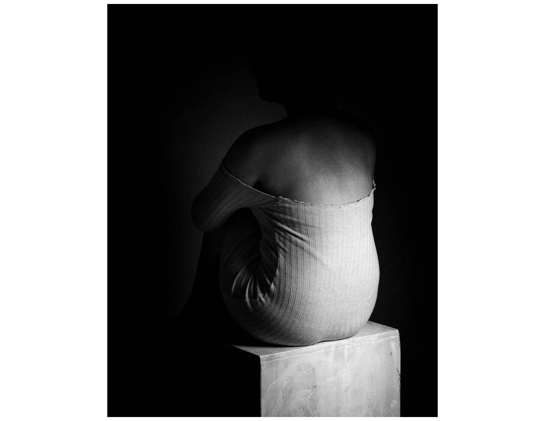 A black and white photograph of a sculpture depicting a human figure with a rounded, smooth body, sitting cross-legged on a box.