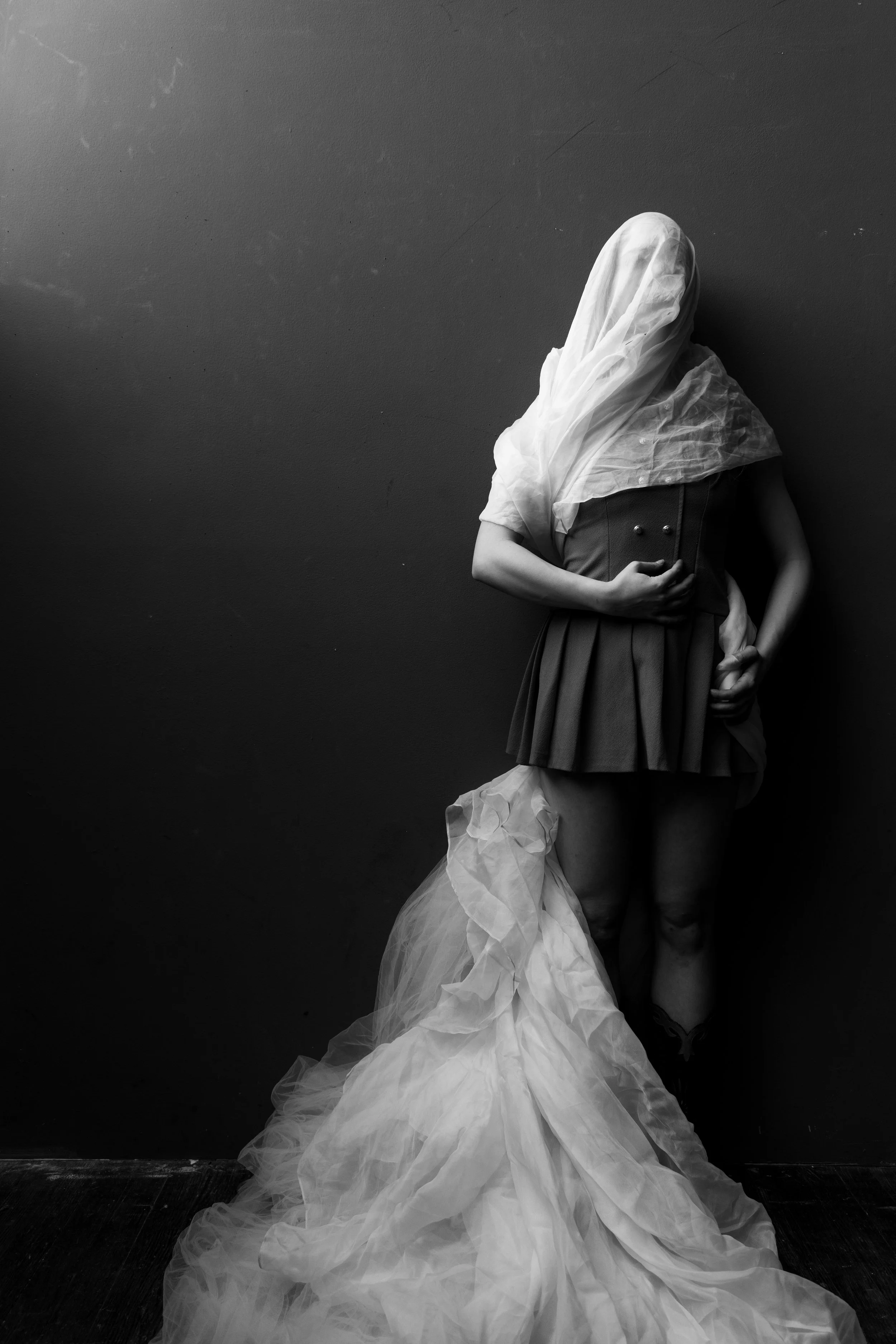 A person standing against a dark wall, wearing a short, pleated skirt, a top, and a veil that covers their head and face, with a long flowing fabric cascading down to the floor.