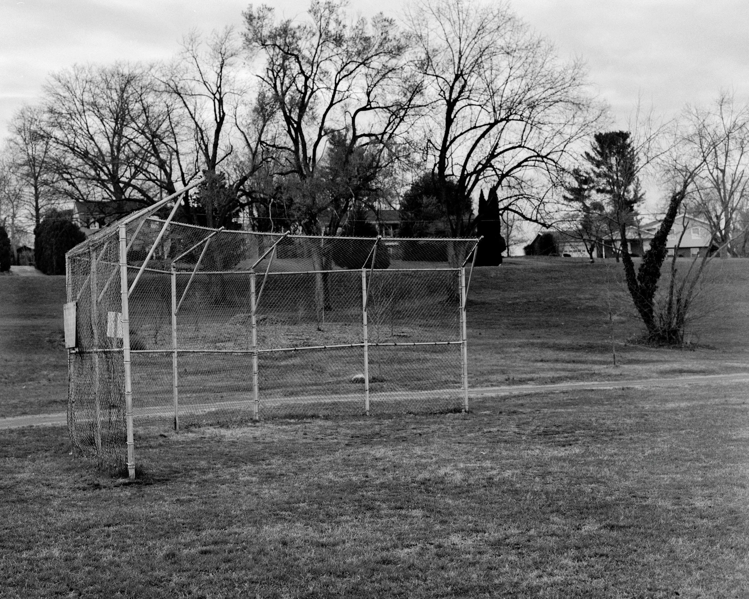 Black and white photo of a baseball field with a damaged fence and leafless trees in the background.