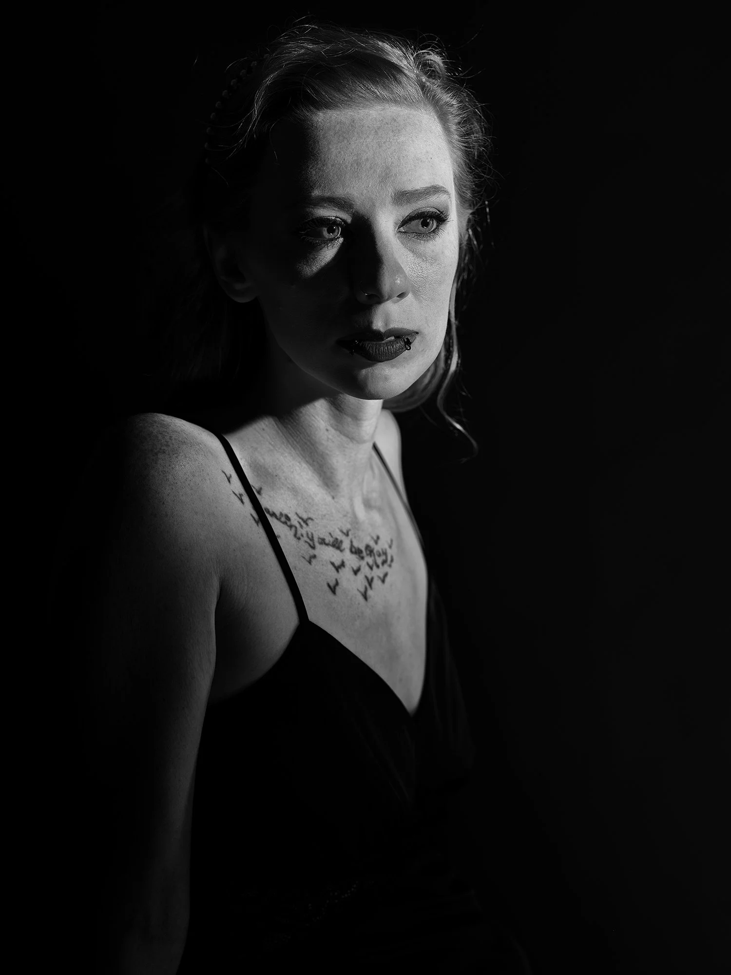 Black and white portrait of a woman with shoulder-length hair, wearing a spaghetti strap top, and tattoos on her chest, looking sideways with dramatic lighting highlighting her face and shoulder.