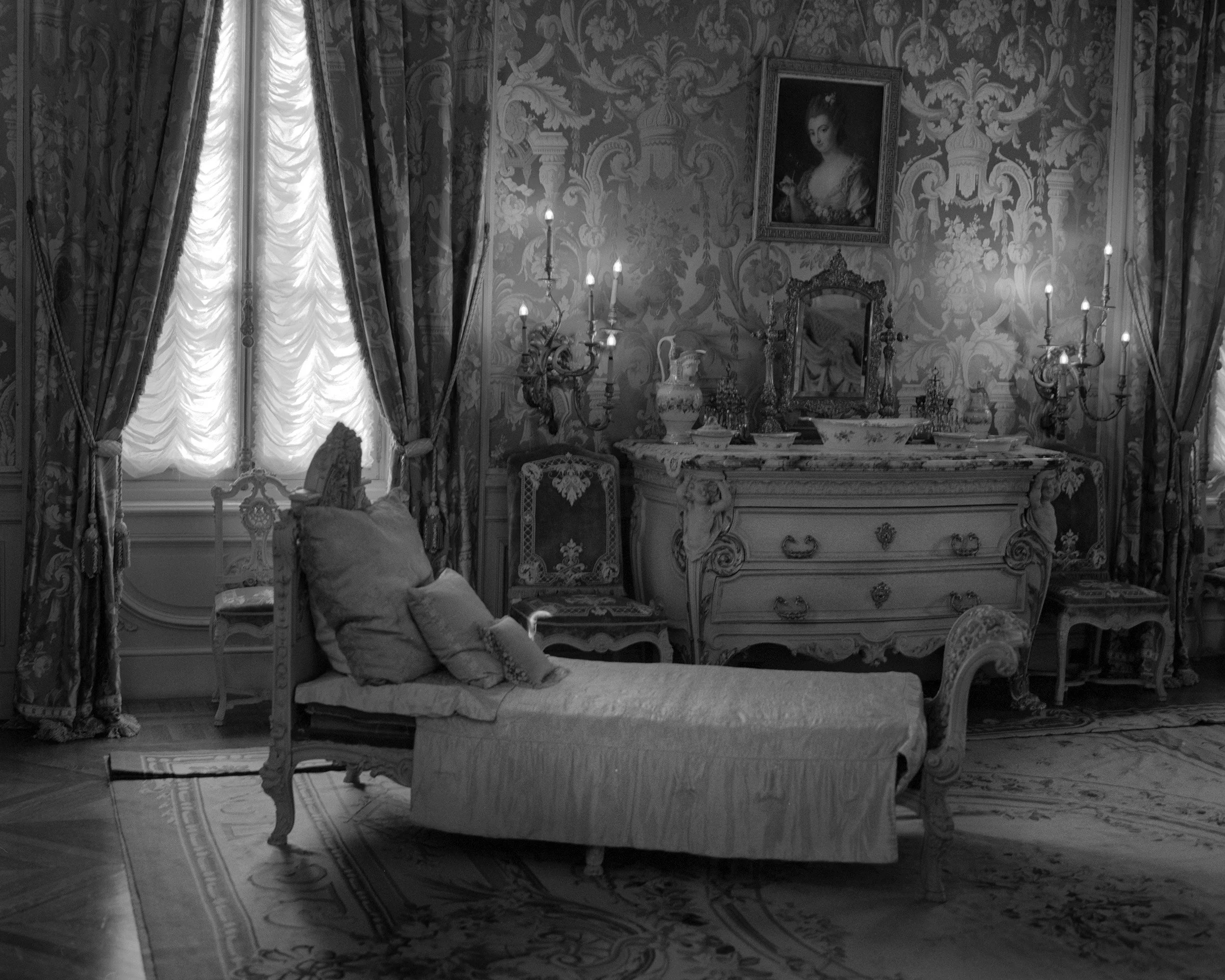 A luxurious bedroom with ornate furniture, patterned wallpaper, a framed portrait on the wall, a large window with elaborate curtains, and a marble-topped dresser with jewelry and decorative items.